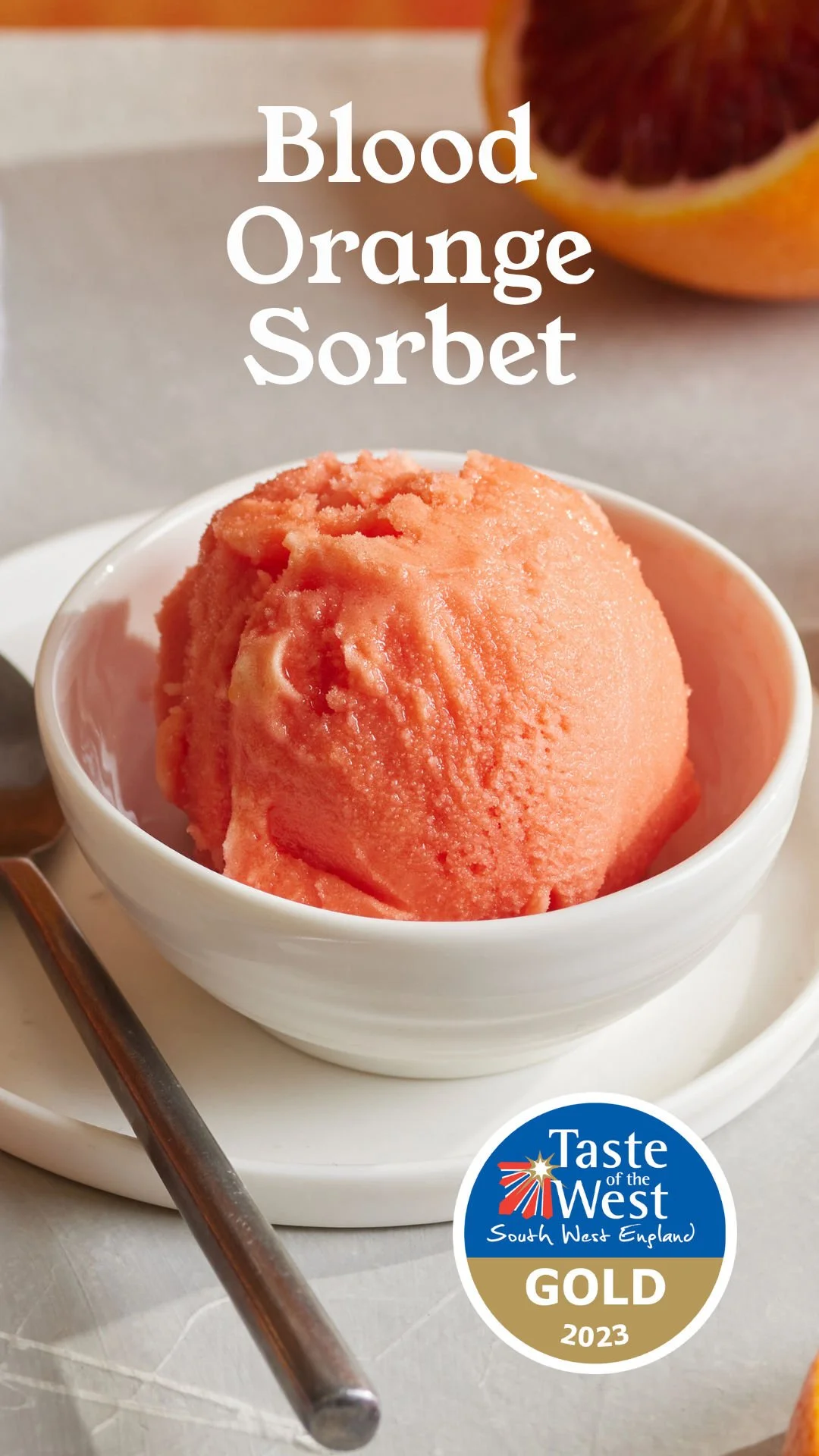 A scoop of blood orange sorbet in a white bowl, with a halved blood orange in the background and a Taste of the West South West England Gold award badge from 2023.