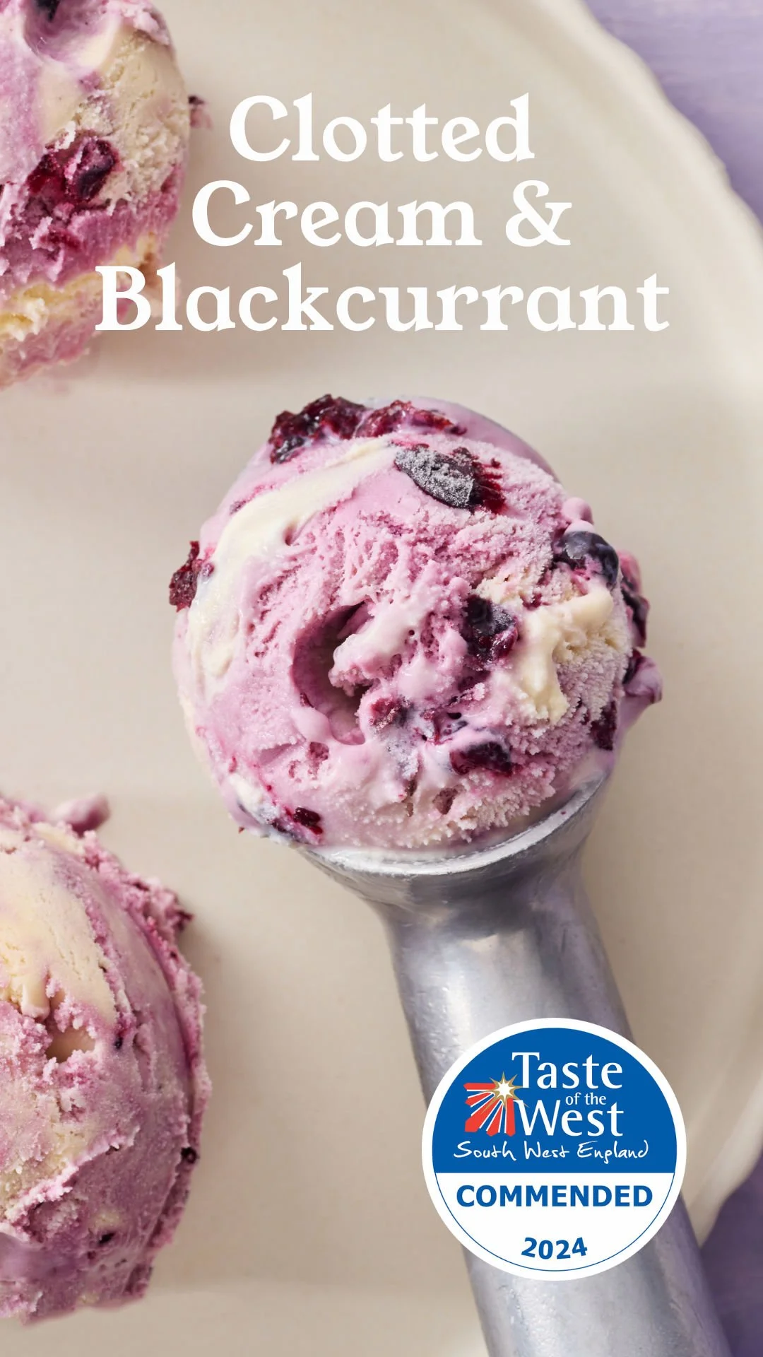 A scoop of pink and white ice cream with blackcurrant pieces in a metal ice cream scoop, with more ice cream scoops partially visible in the background and a circular blue and white emblem with text at the bottom right.