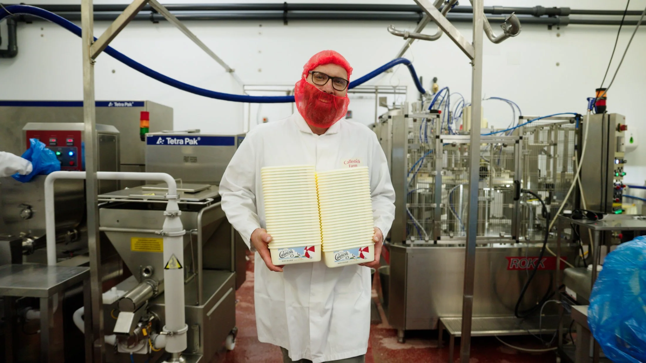 A man in white lab coat with red beard and glasses holding six cartons of cheese in a dairy processing plant.