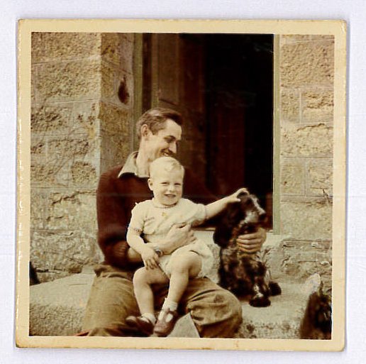 A vintage photo of a man with a young child and a small dog sitting outdoors in front of a stone building.