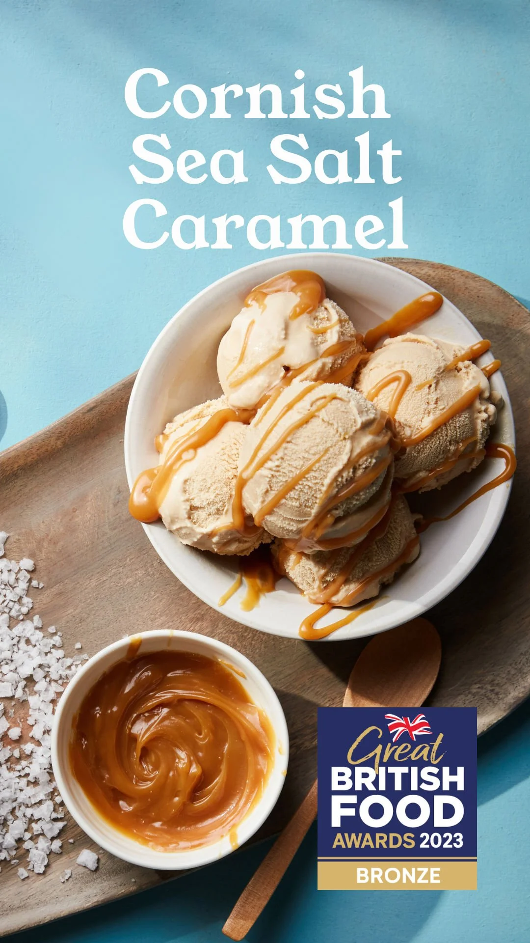 Scoops of caramel ice cream drizzled with caramel sauce on a white bowl, with a small bowl of caramel sauce and sea salt on a wooden tray.
