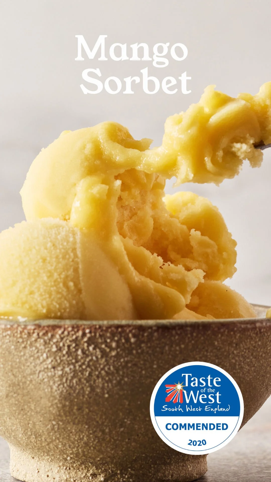 Close-up of a bowl filled with creamy yellow mango sorbet, with a spoon lifting a scoop. The bowl has a textured, speckled surface. There's a Taste of the West badge on the bowl indicating it was commended in 2020.