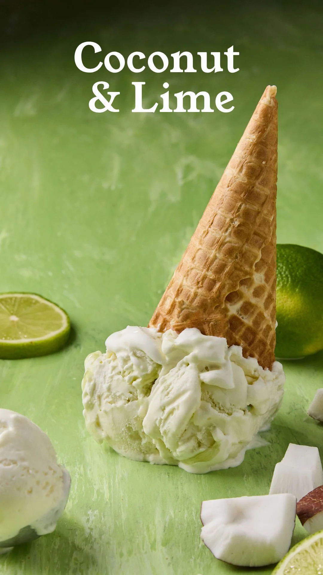 A scoop of coconut and lime flavored ice cream with an upside-down waffle cone on top, surrounded by lime slices and coconut pieces on a green background.