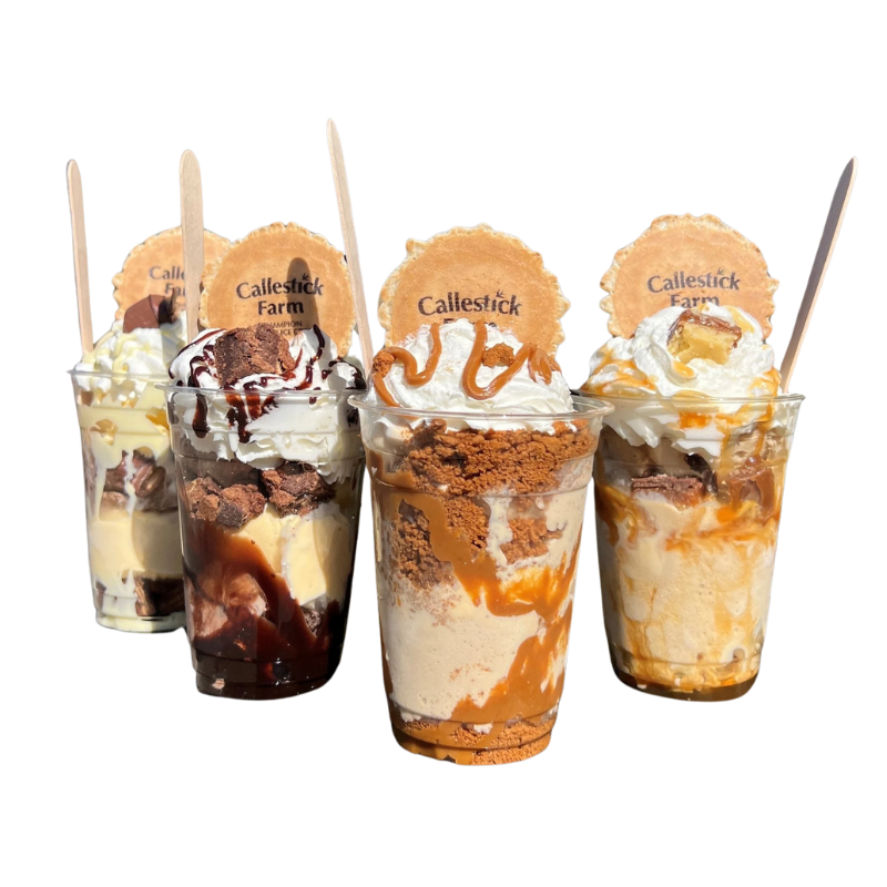 Four large dessert parfaits containing ice cream, whipped cream, chocolate and caramel sauces, cookie pieces, and biscuit toppings, each labeled with a 'Callestick Farm' cookie, against a black background.