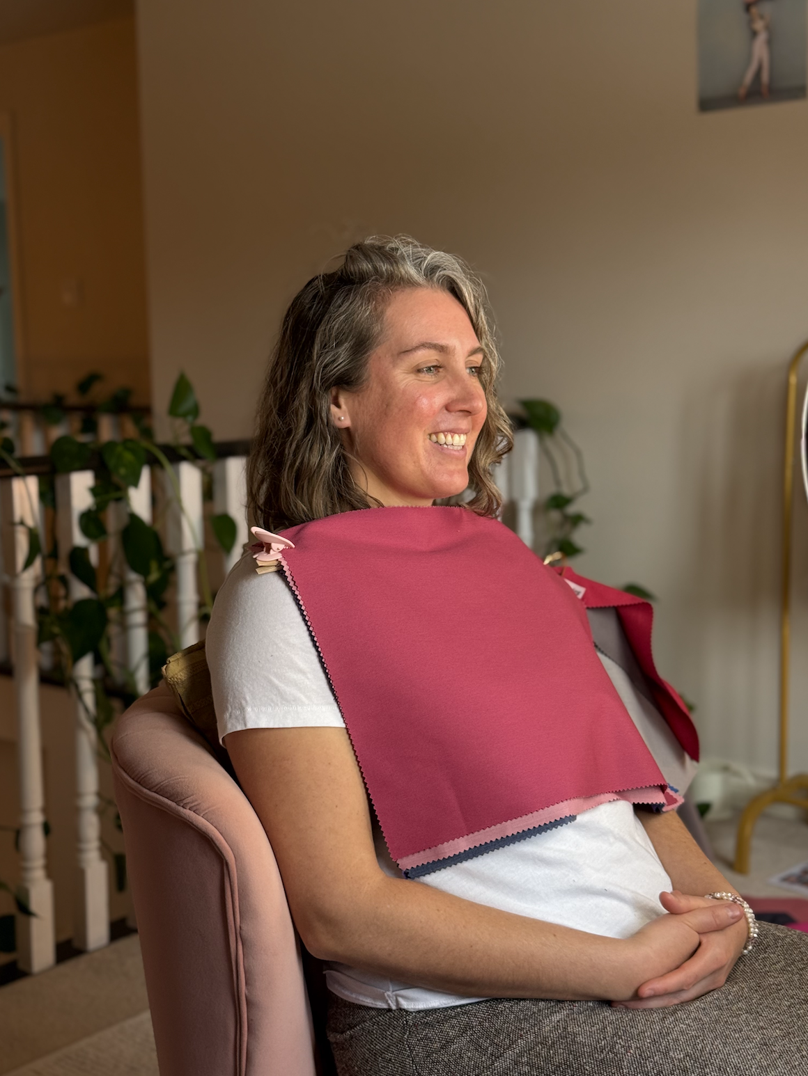 A woman with shoulder-length wavy gray hair sitting in a pink chair, smiling, wearing a white t-shirt and a red bib draped over her shoulders.