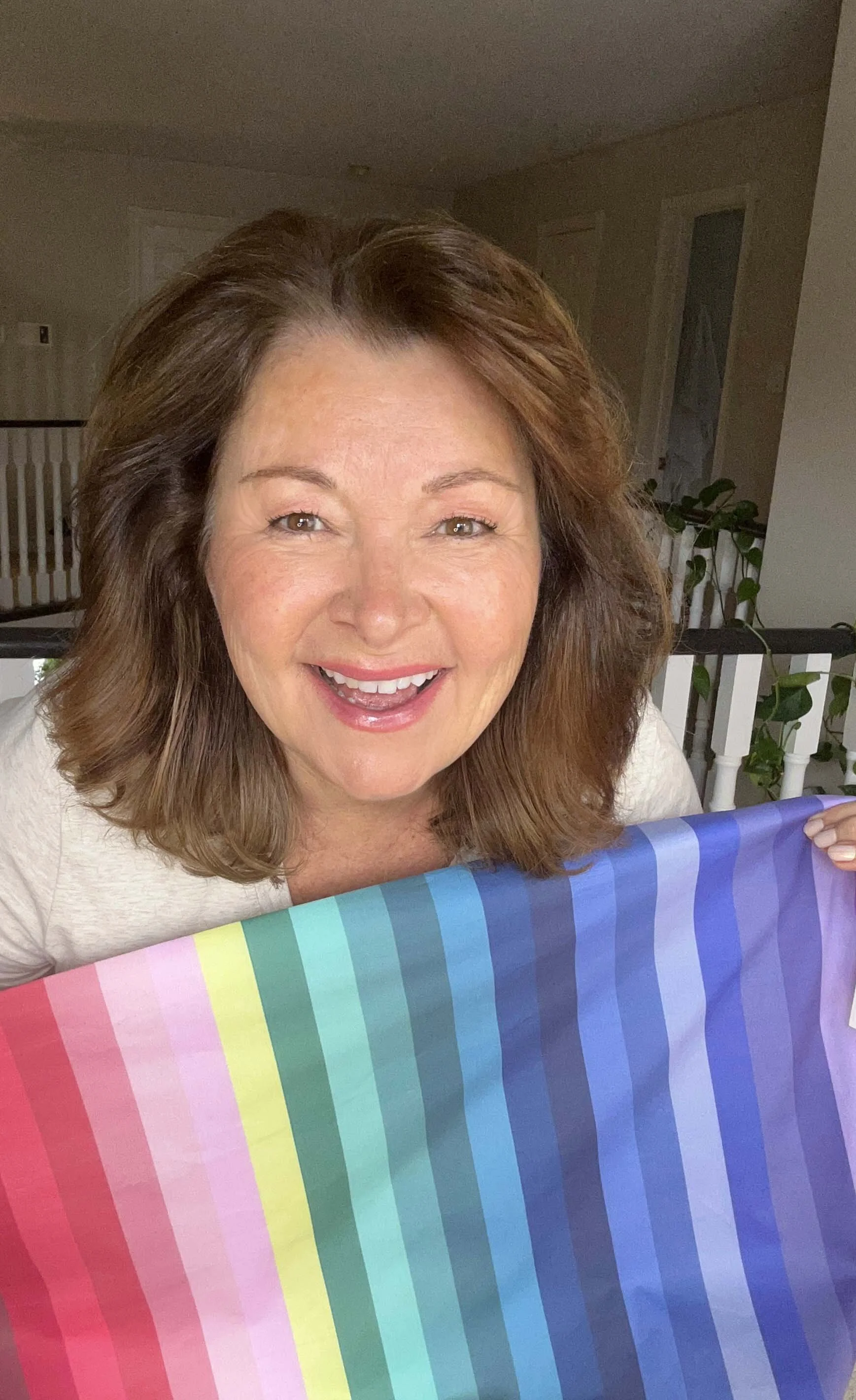 A smiling middle-aged woman with brown hair, holding a rainbow-colored Pride flag. She is indoors, with a railing and some plants in the background.