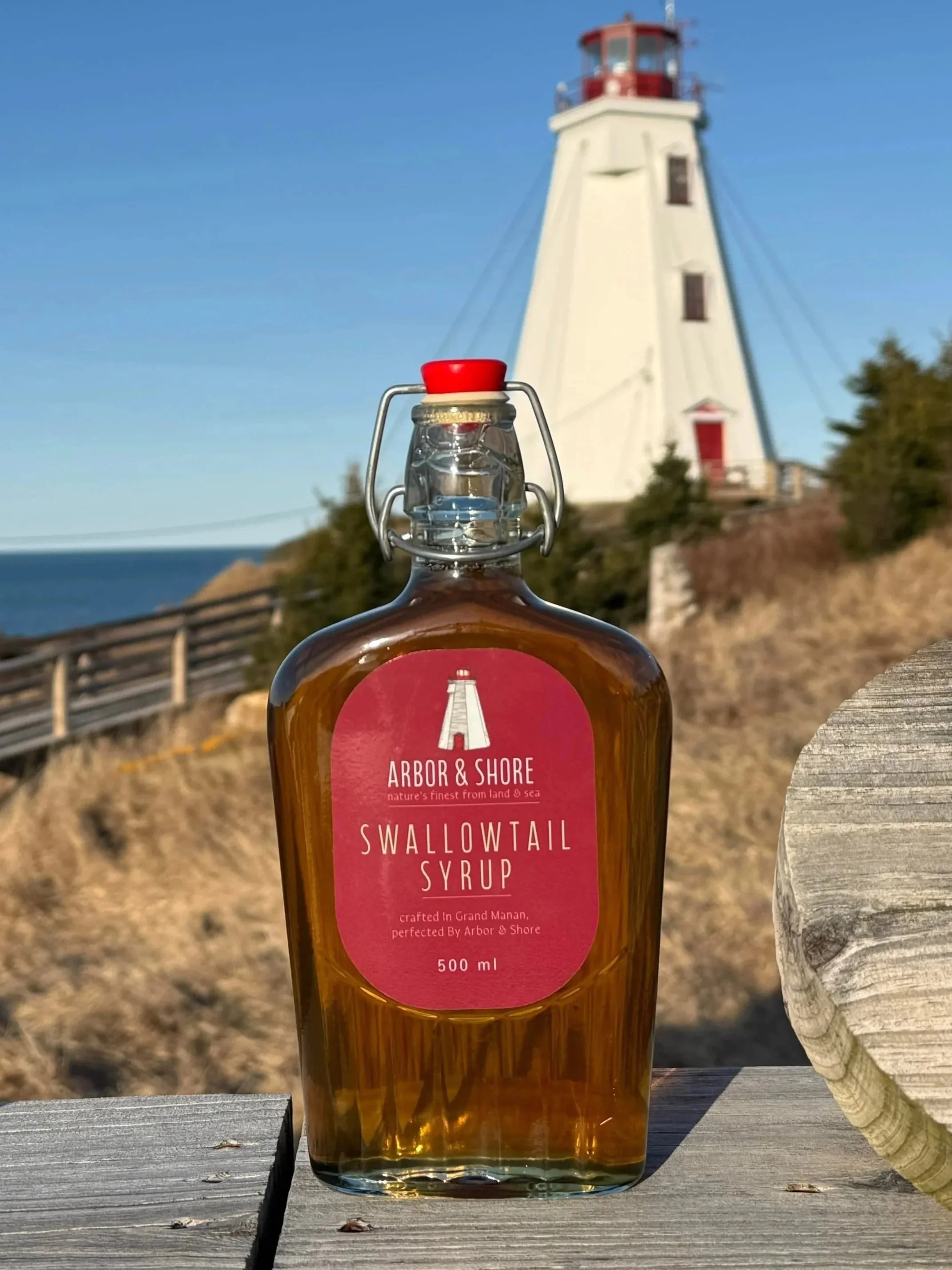 A bottle of Arbor & Shore Swallowtail Syrup placed on a wooden surface outdoors, with a lighthouse in the background on a clear, sunny day.
