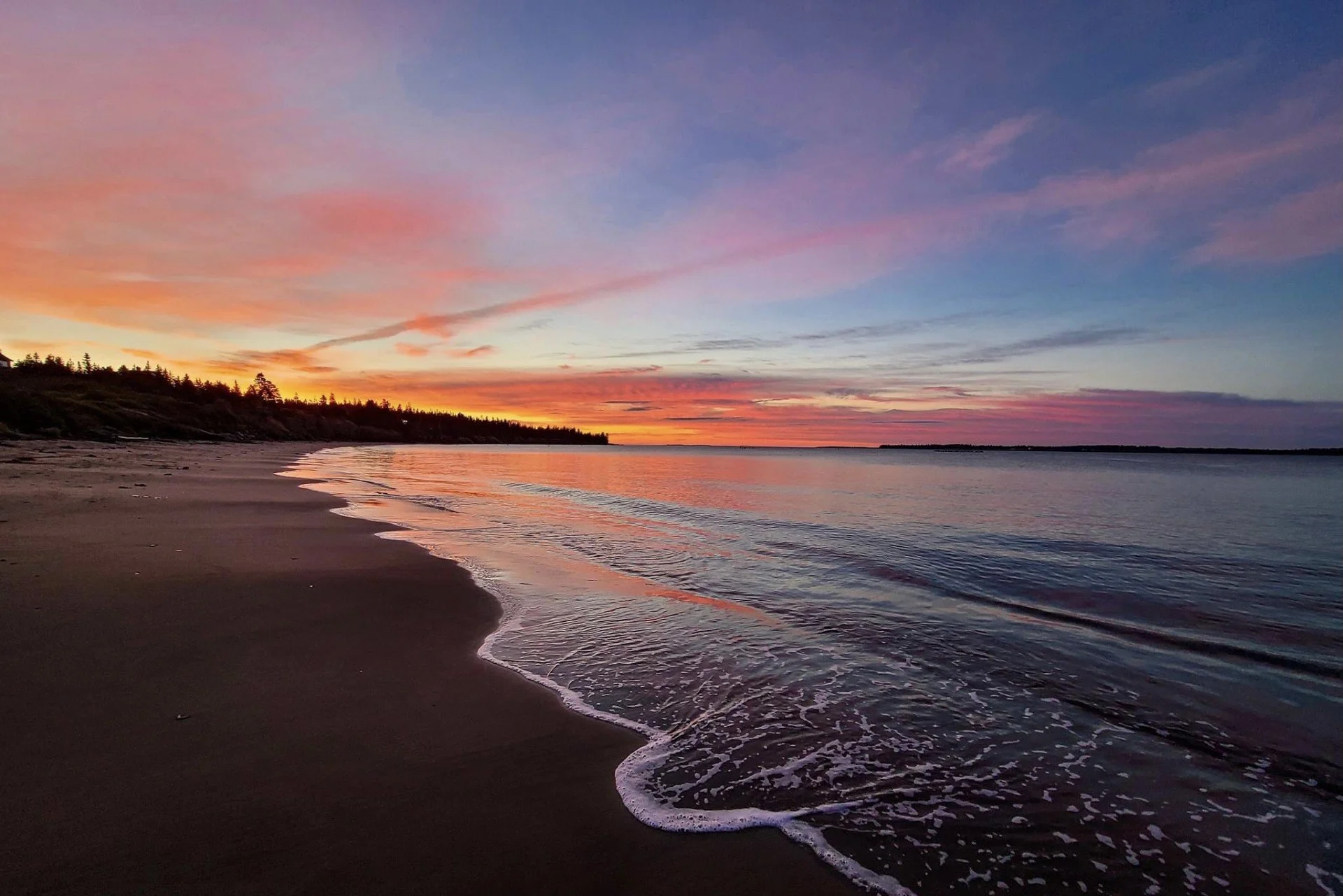 Sunset over a calm beach with colorful sky, gentle waves, and trees on the shoreline.