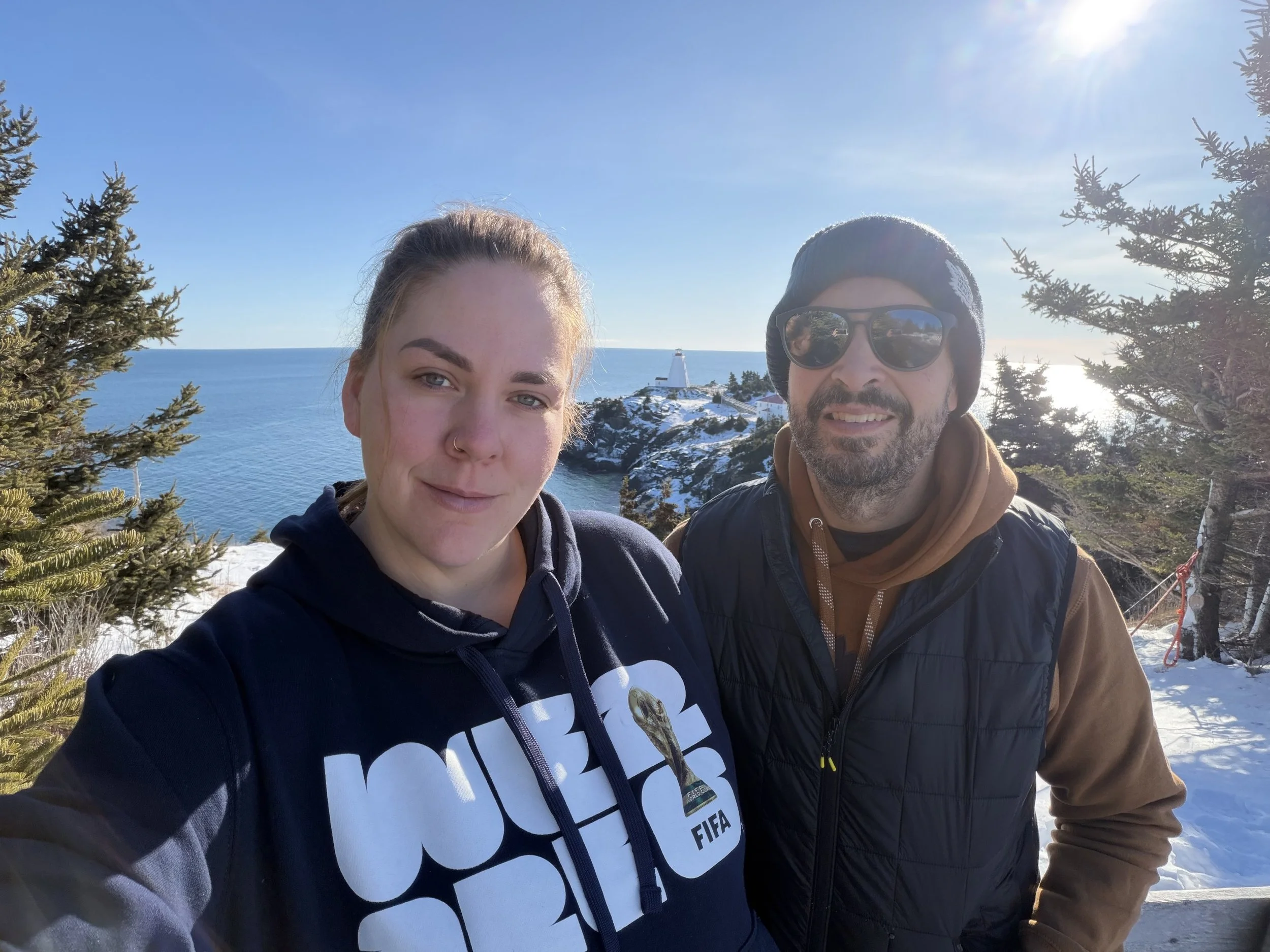 A young woman and a man posing for a selfie outdoors in winter, with snow, trees, and a lighthouse visible on a rocky outcrop over the ocean in the background on Grand Manan Island