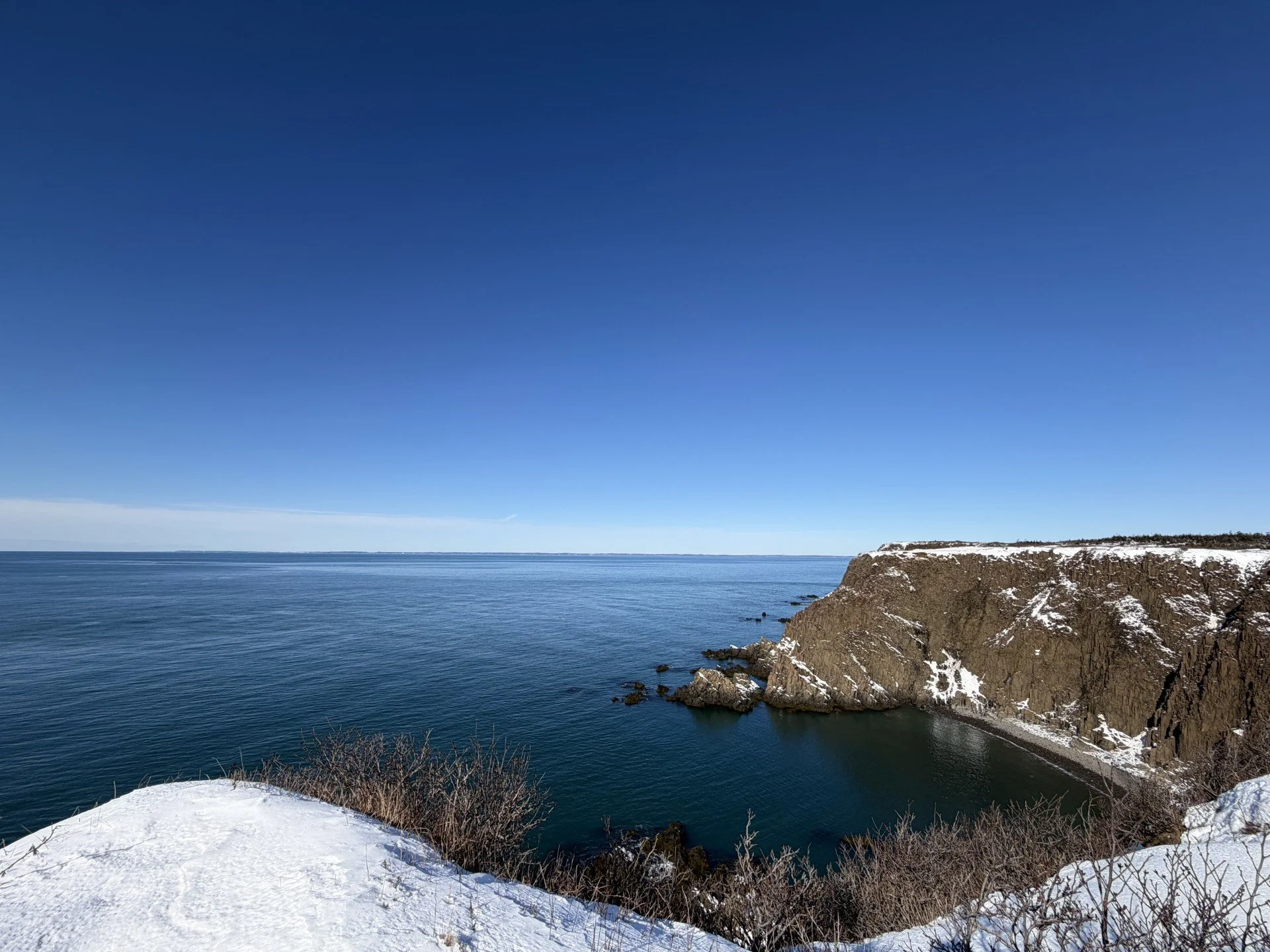 Snow-covered coastline with a rocky cliff and calm ocean under a clear blue sky.