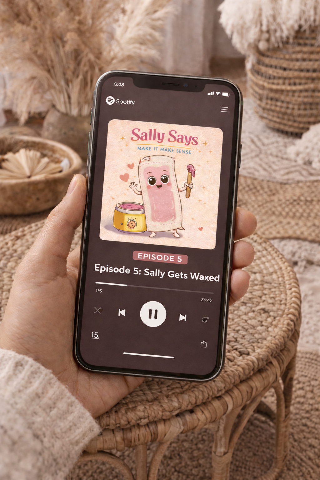 A person holding a smartphone displaying a Spotify podcast episode titled 'Episode 5: Sally Gets Waxed' from the series 'Sally Says' with an animated cartoon of a character named Sally holding a wax applicator, with a wax pot nearby.