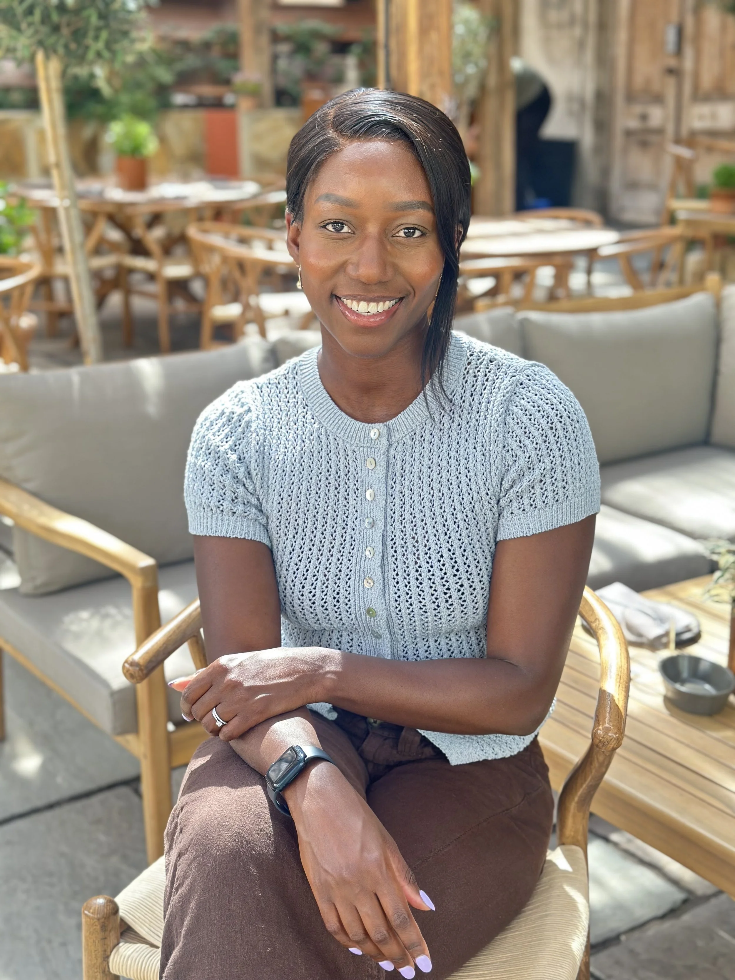 A smiling woman with short dark hair sitting in a wooden outdoor seating area with plants and rustic decor.