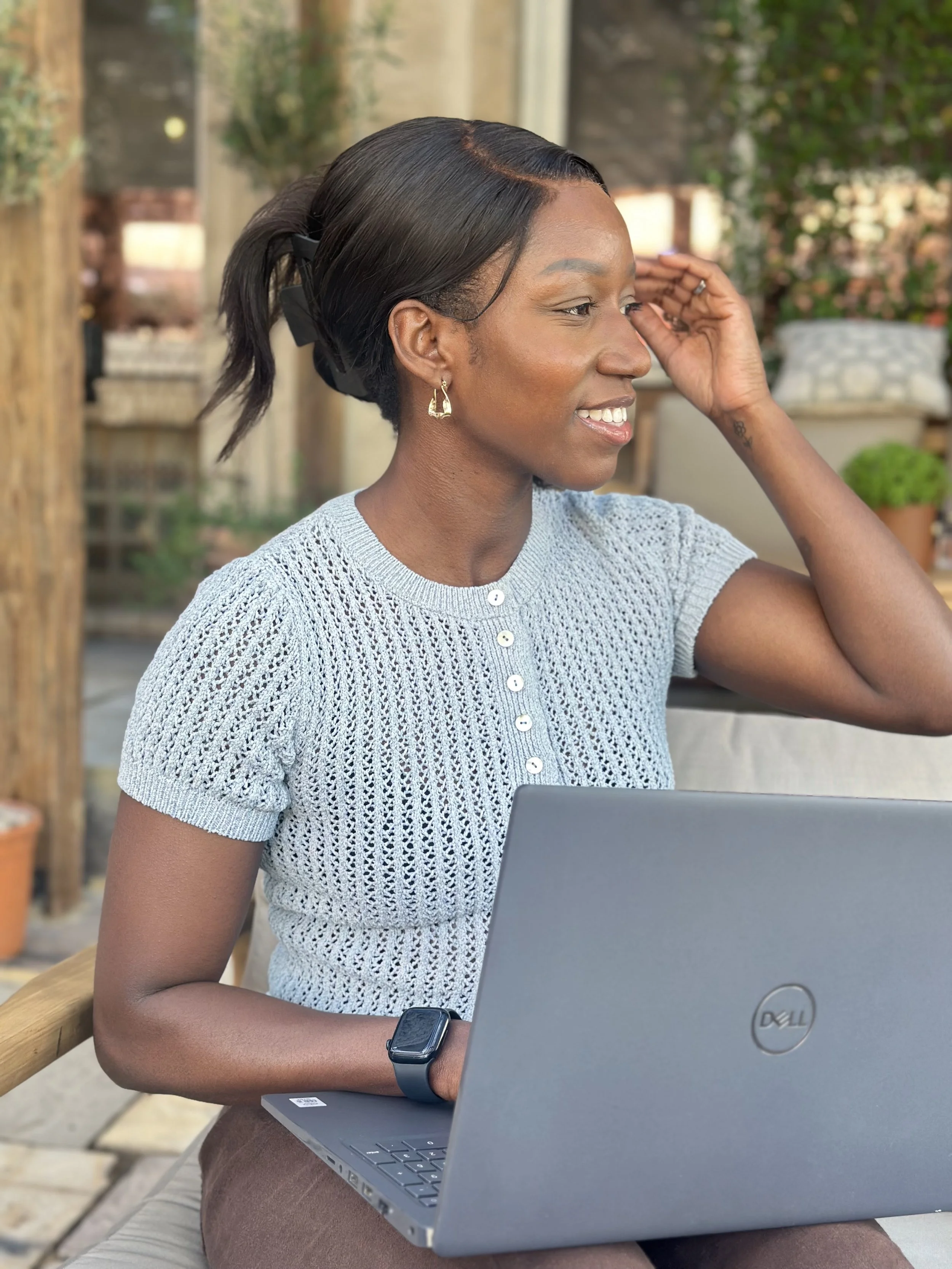 A woman sitting outdoors at a wooden table using a Dell laptop, wearing a knitted gray top, black smartwatch, earrings, and a ring, with trees and plants in the background.