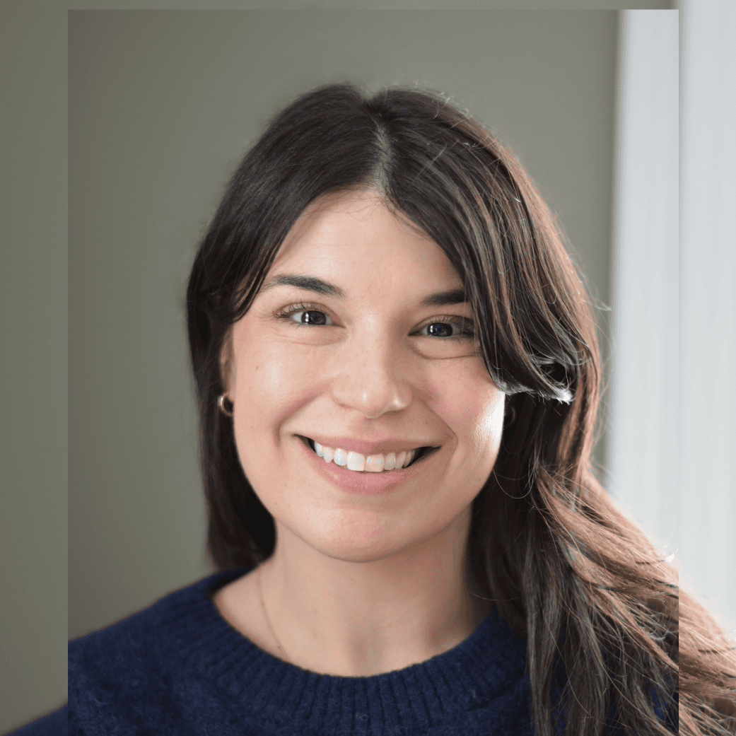 A woman with long dark hair smiling, wearing a navy sweater, indoors near a window.