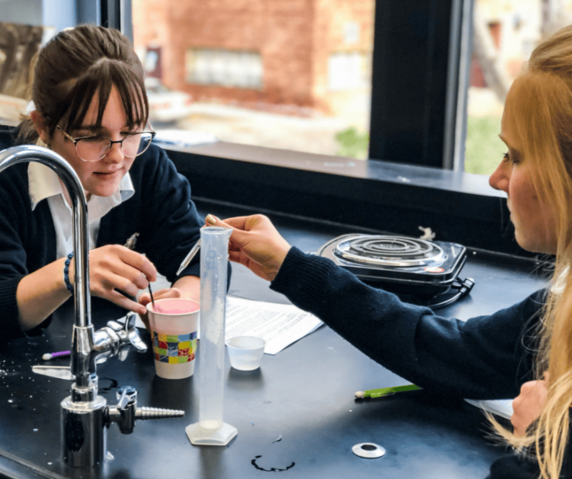 Two young women conducting a science experiment at a lab table, with one of them holding a pipette over a cup filled with pink liquid.