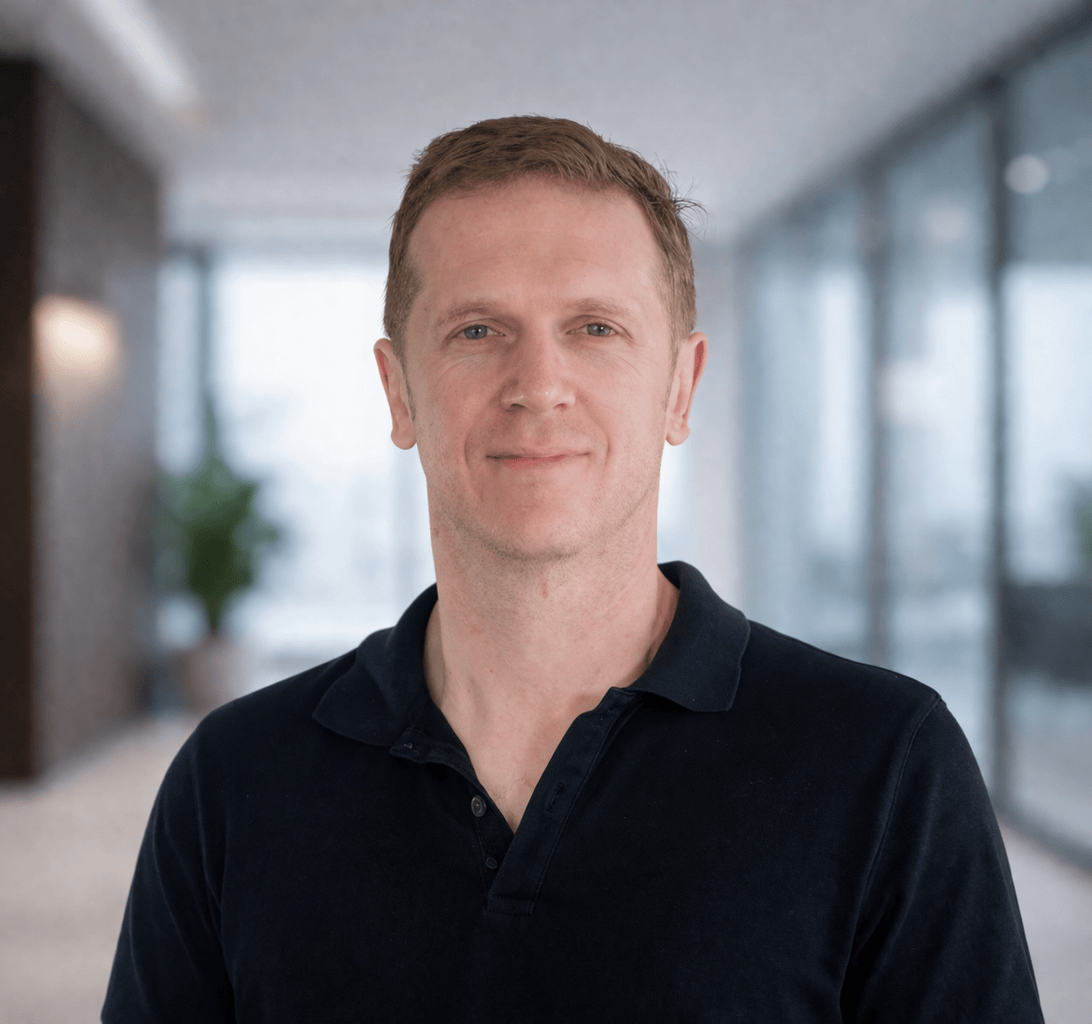Portrait of a man with short light brown hair and fair skin, wearing a dark collared shirt, standing in a modern indoor space with large windows and blurred background.