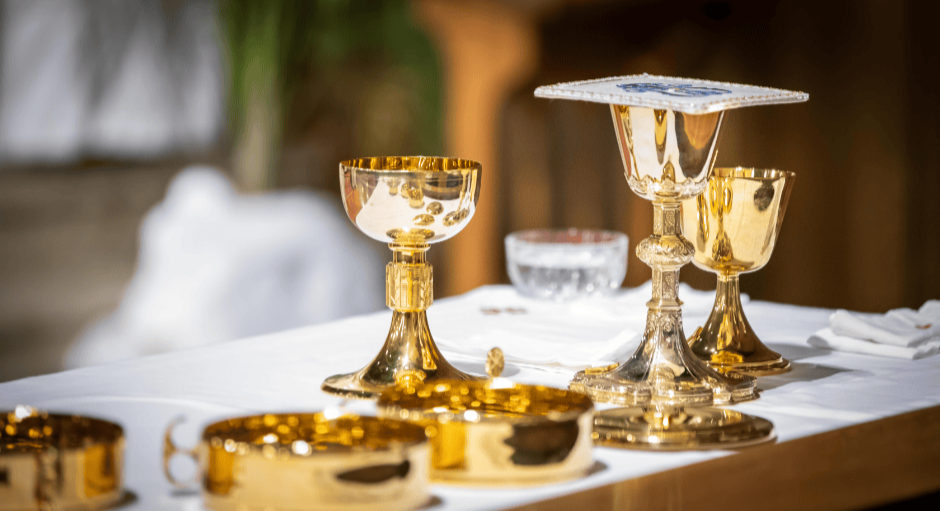 Golden goblets and chalices on a white tablecloth, with some silver dishes and a deck of cards, in a cozy indoor setting.