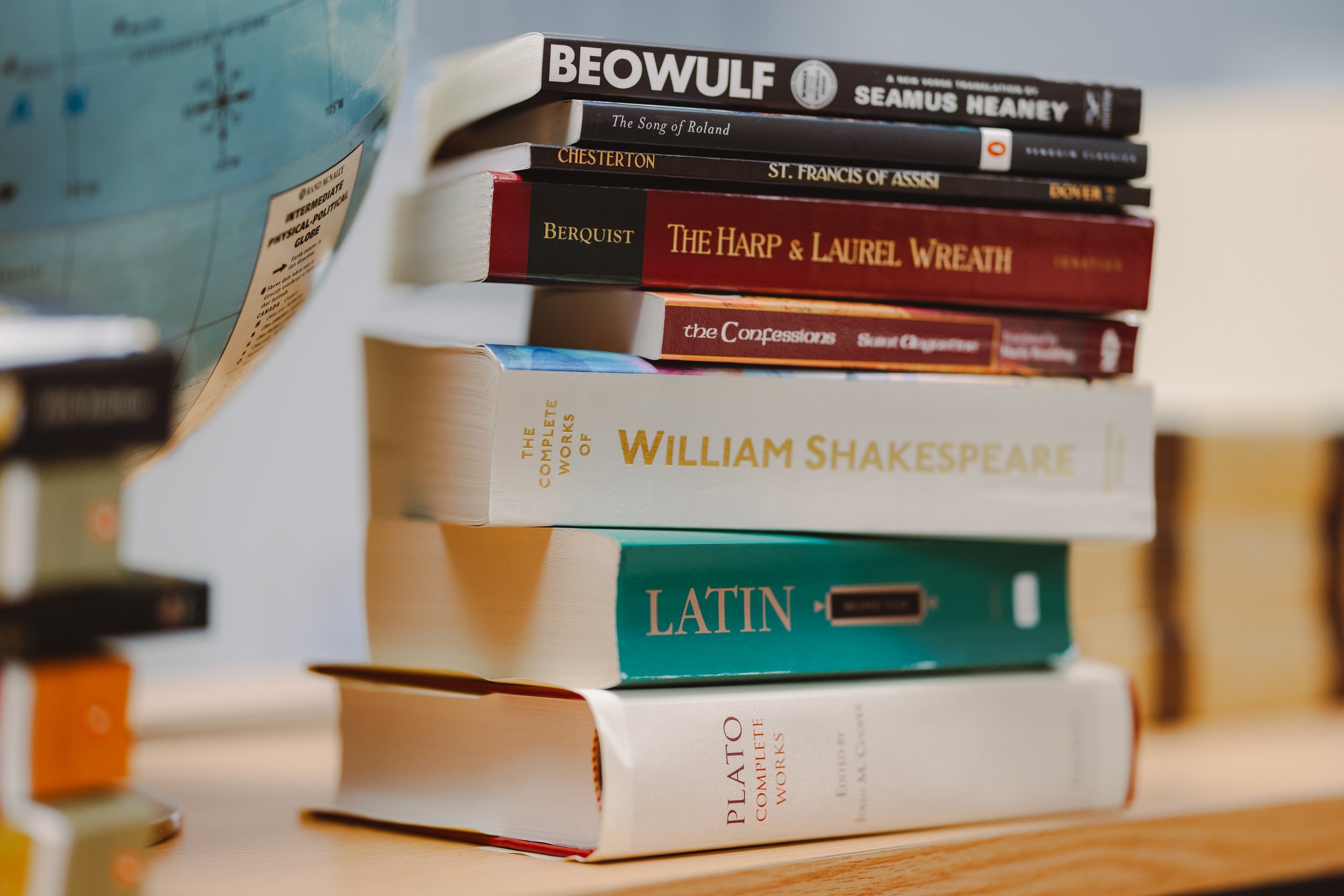 Stack of several books including works by William Shakespeare, Latin, Plato, and others, placed on a wooden surface next to a globe.