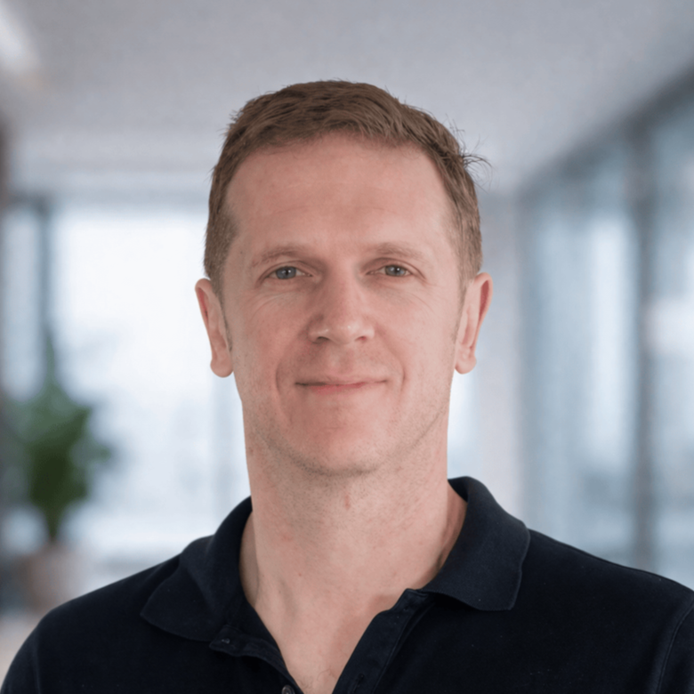 Headshot of a man with short, light brown hair, wearing a black collared shirt, smiling gently, in an office setting with blurred background.