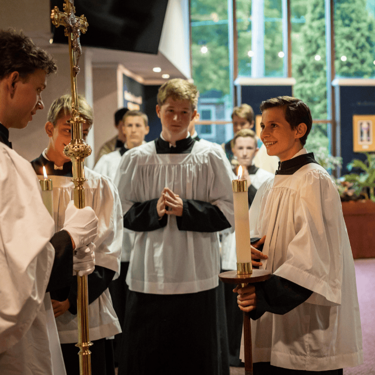 Young boys dressed in choir robes gathered around a priest during a religious ceremony, inside a church with large windows and greenery outside.