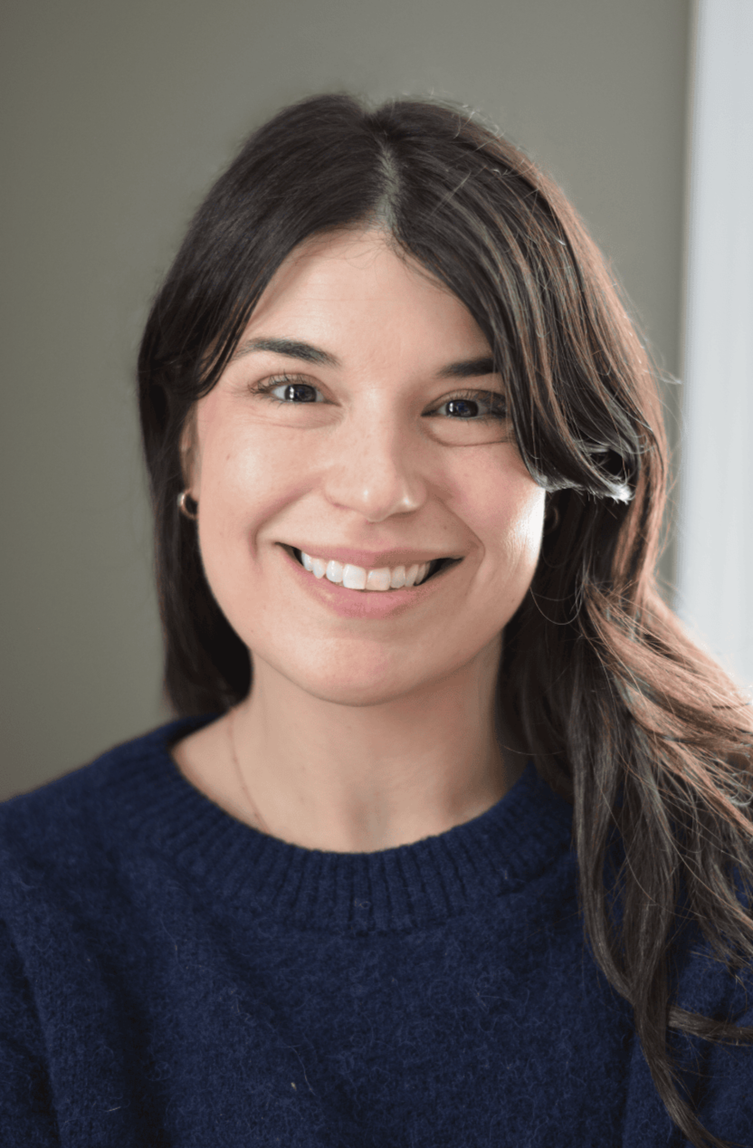 A young woman with shoulder-length dark brown hair smiling, wearing a navy blue sweater indoors.