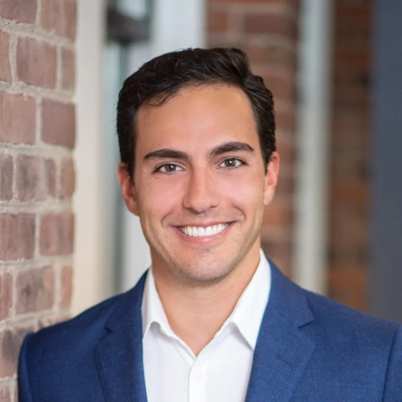 Headshot of a smiling man in a navy blazer and white shirt, standing outdoors next to a brick wall.