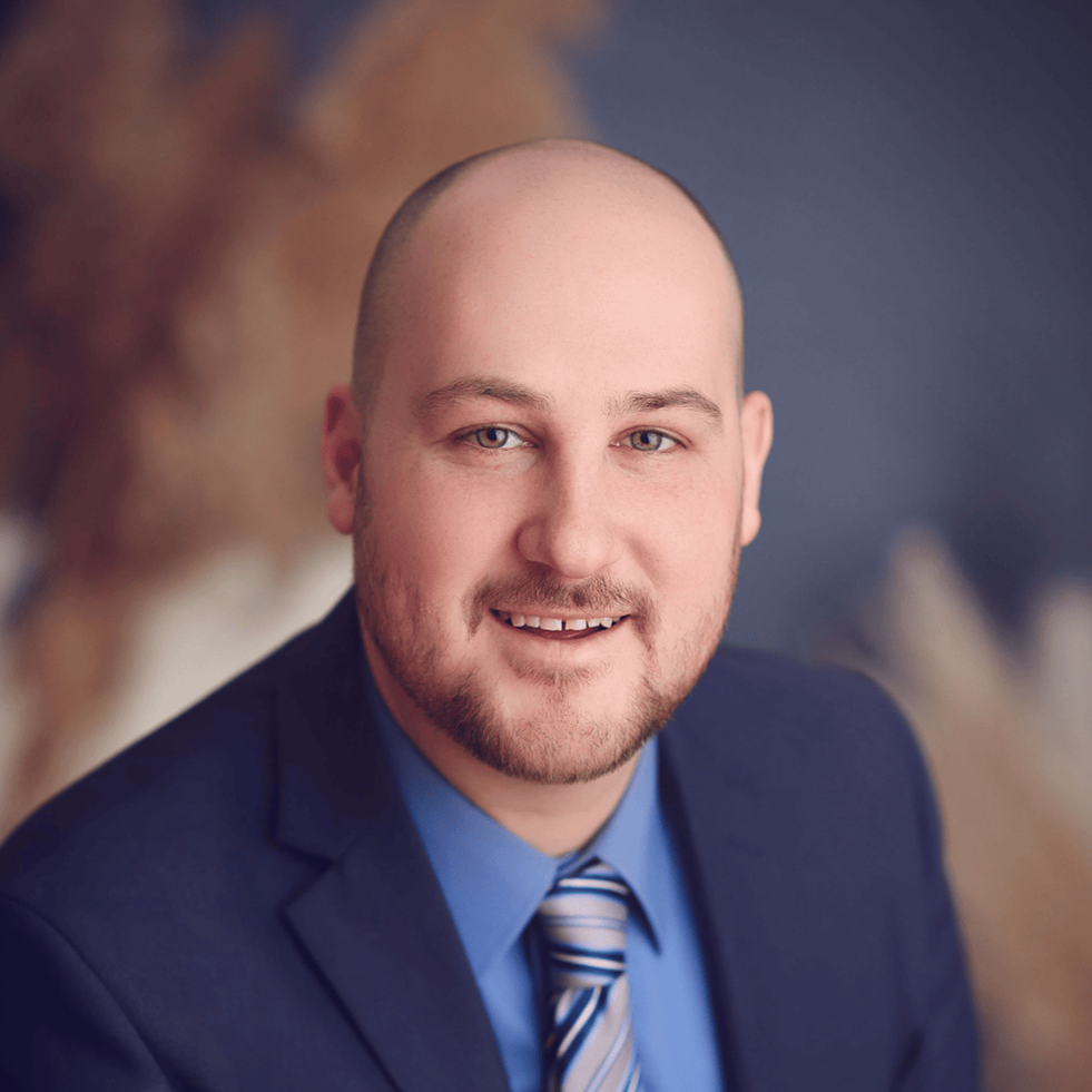 Professional headshot of a man in a suit and tie, with a blurred autumnal background.