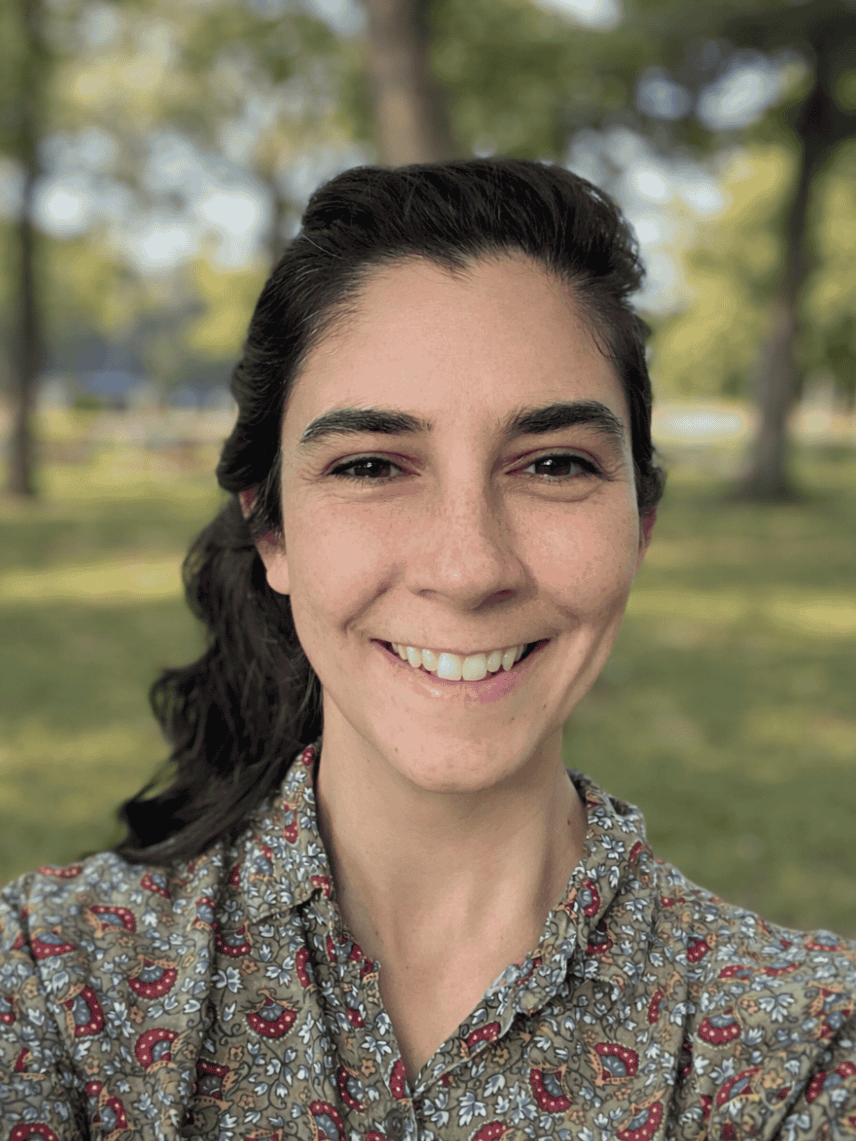 A smiling woman with dark hair tied back, wearing a patterned shirt, outdoors with trees and grass in the background.