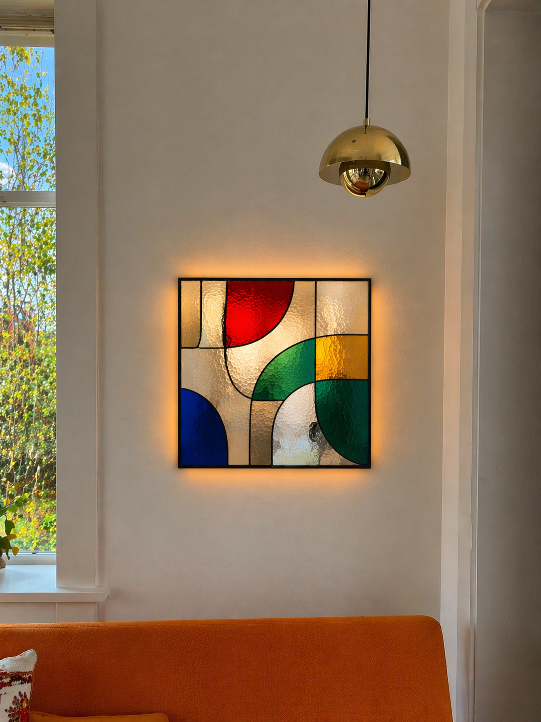 Kunstwerk met gekleurde geometrische vormen op een muur met een achtergrondverlichting, boven een oranje bank en naast een raam met buitenzicht.