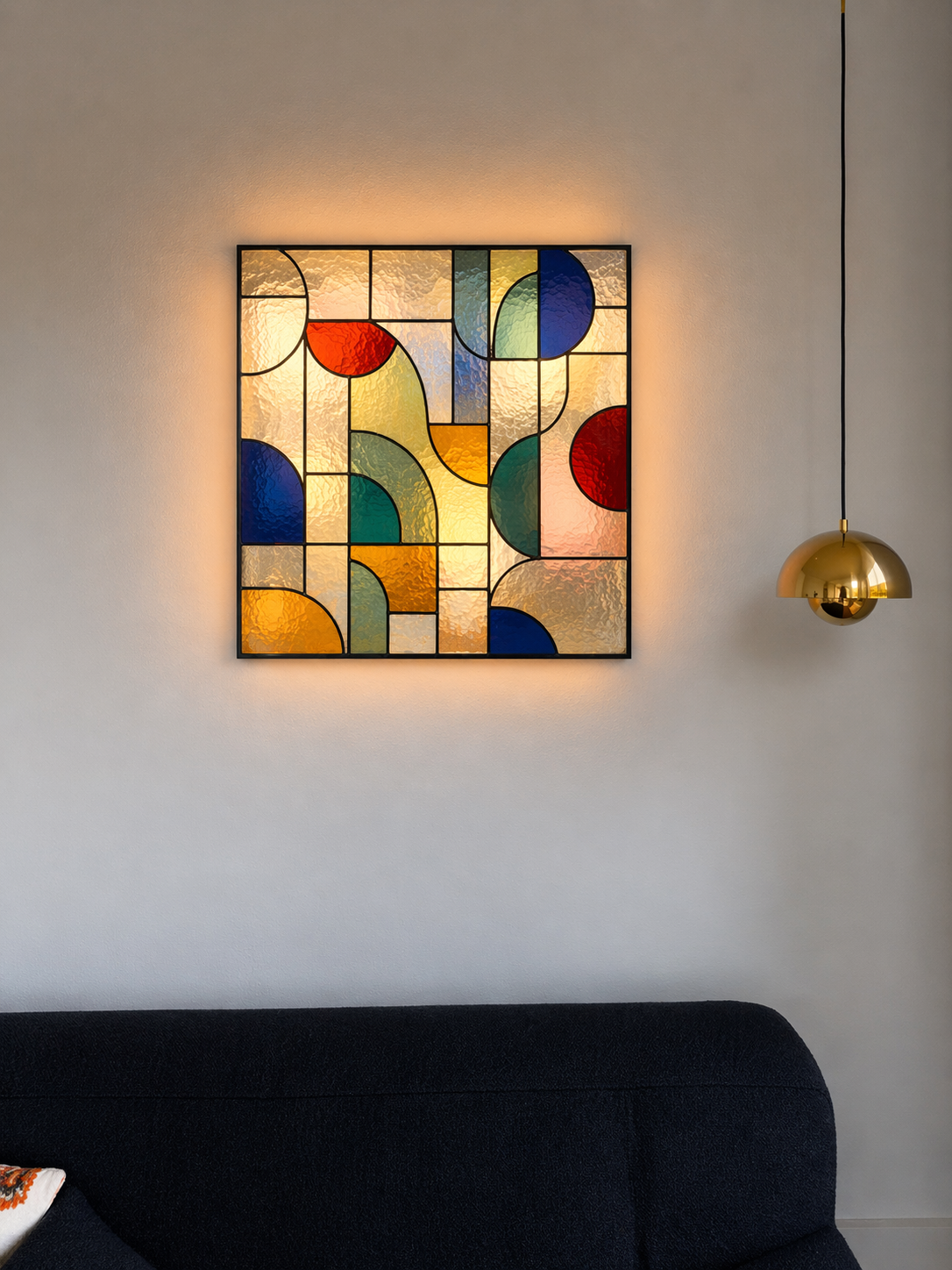 Kleurrijk stained glass kunstwerk met geometrische patronen en achterlicht, hangend aan een wit muur naast een gouden hanglamp.