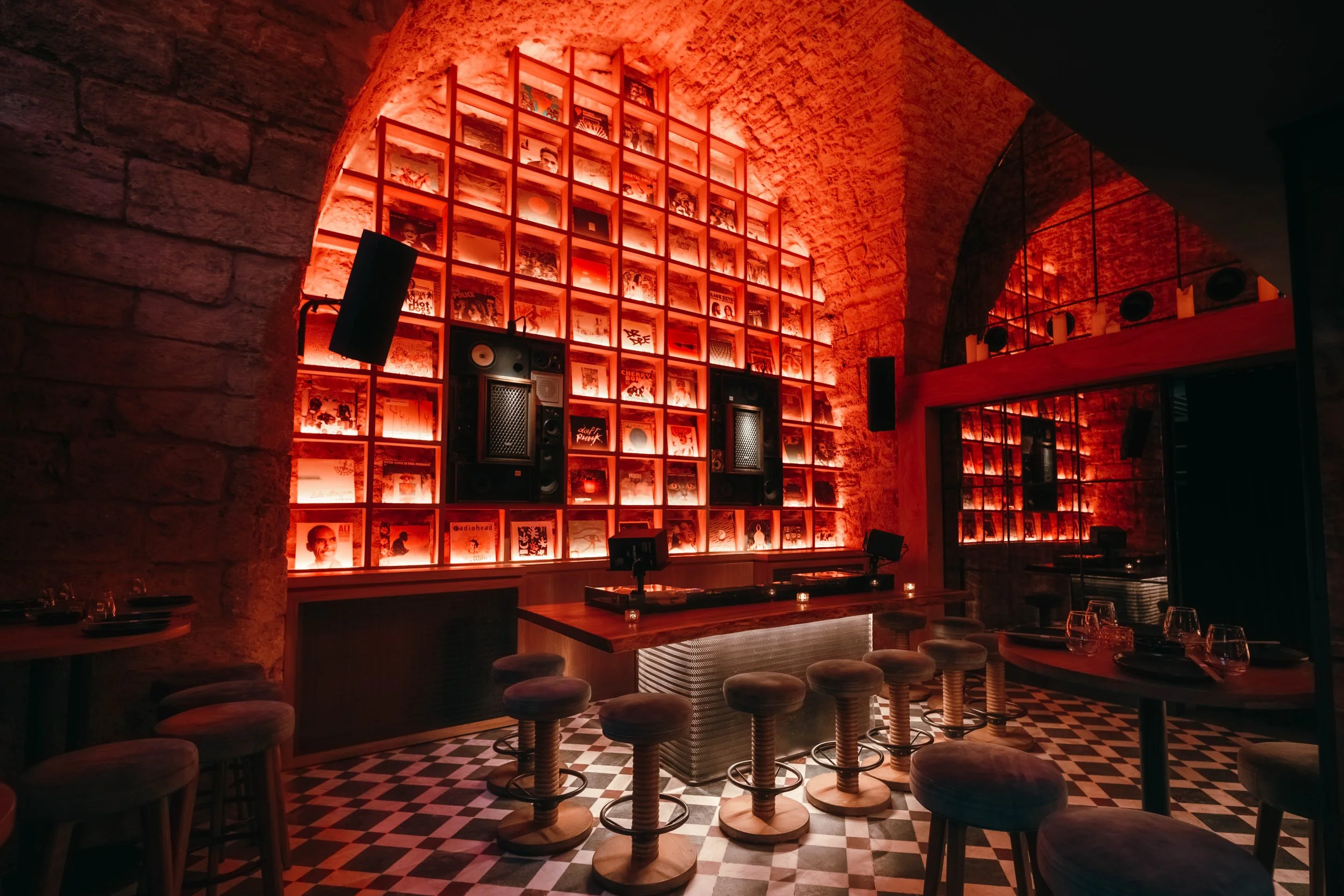 A dimly lit bar or lounge with red lighting, brick walls, a high arched ceiling, shelves filled with vinyl records, a DJ booth with turntables, and barstools around the counter.