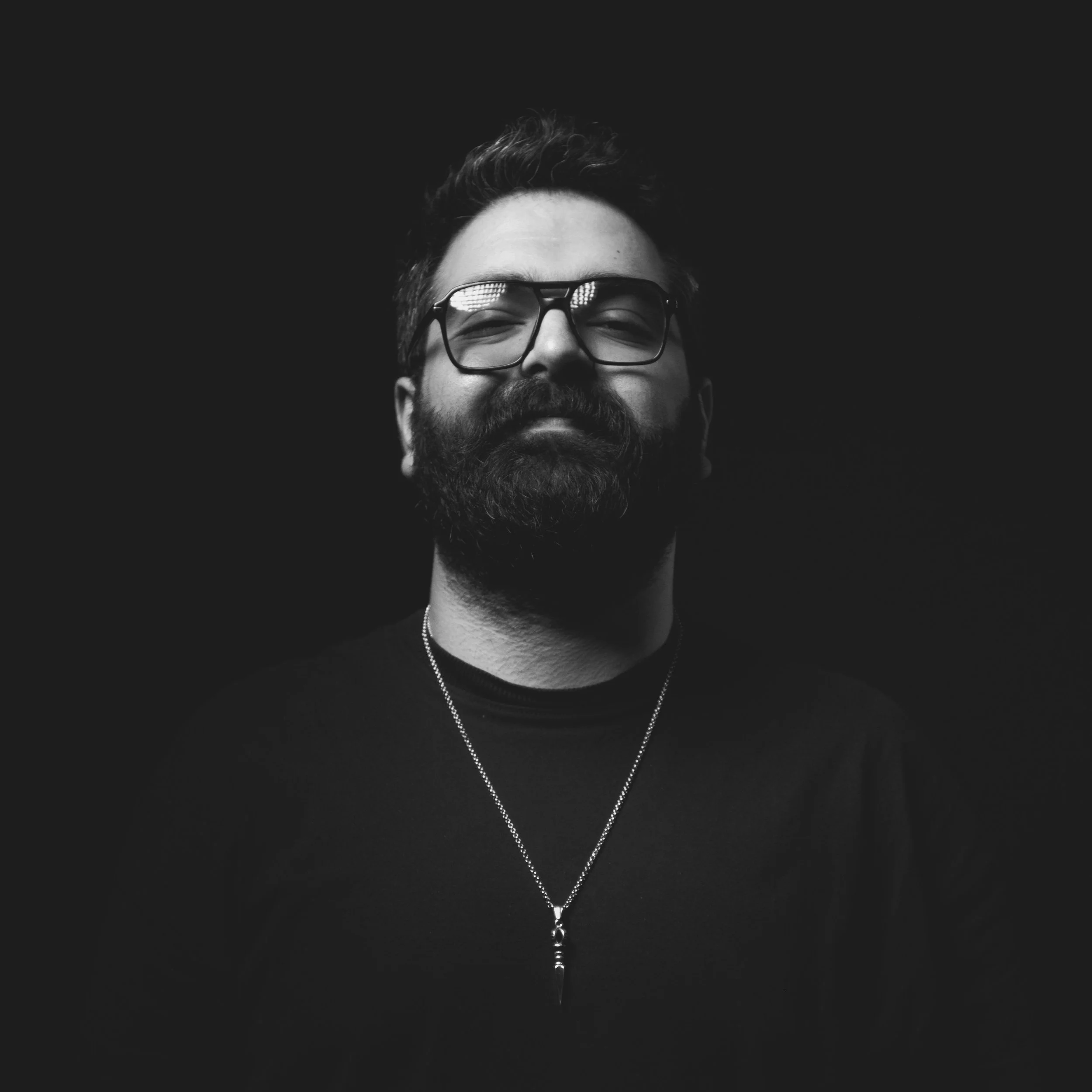 Black and white photo of a bearded man with glasses, smiling slightly, wearing a necklaces and a black shirt, against a dark background.