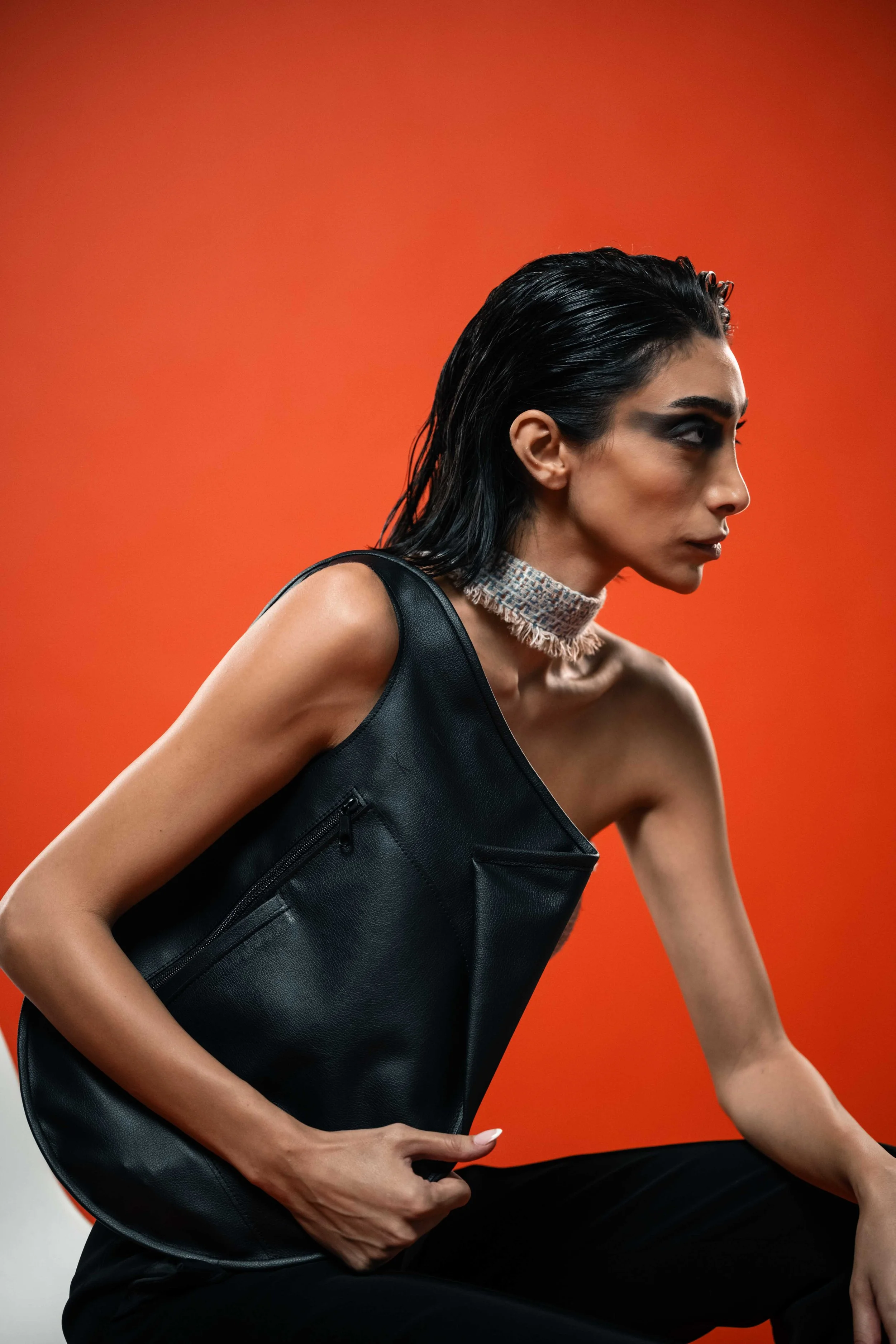 A woman with dark, wet hair styled back, wearing a sleeveless black leather top, a textured choker, and dark makeup, sitting against an orange background.