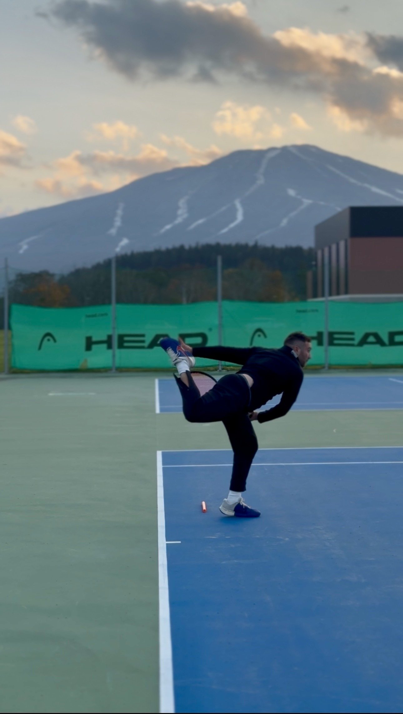 A person dressed in black, wearing a mask, standing on one leg on a tennis court, holding their other leg behind them with one hand, with mountains and a cloudy sky in the background.