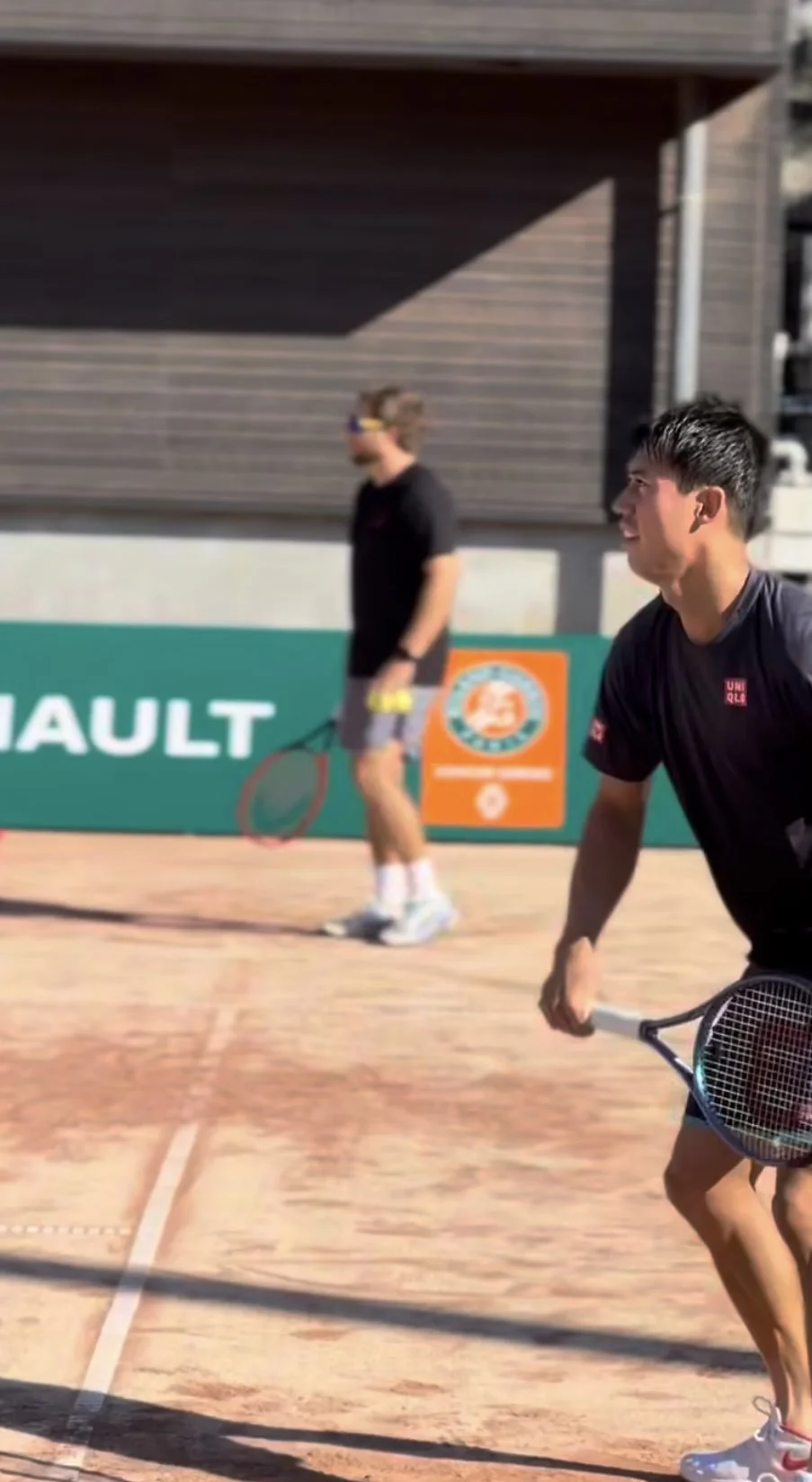 Two male tennis players holding rackets on a tennis court, with one in the foreground and one in the background, during a match.
