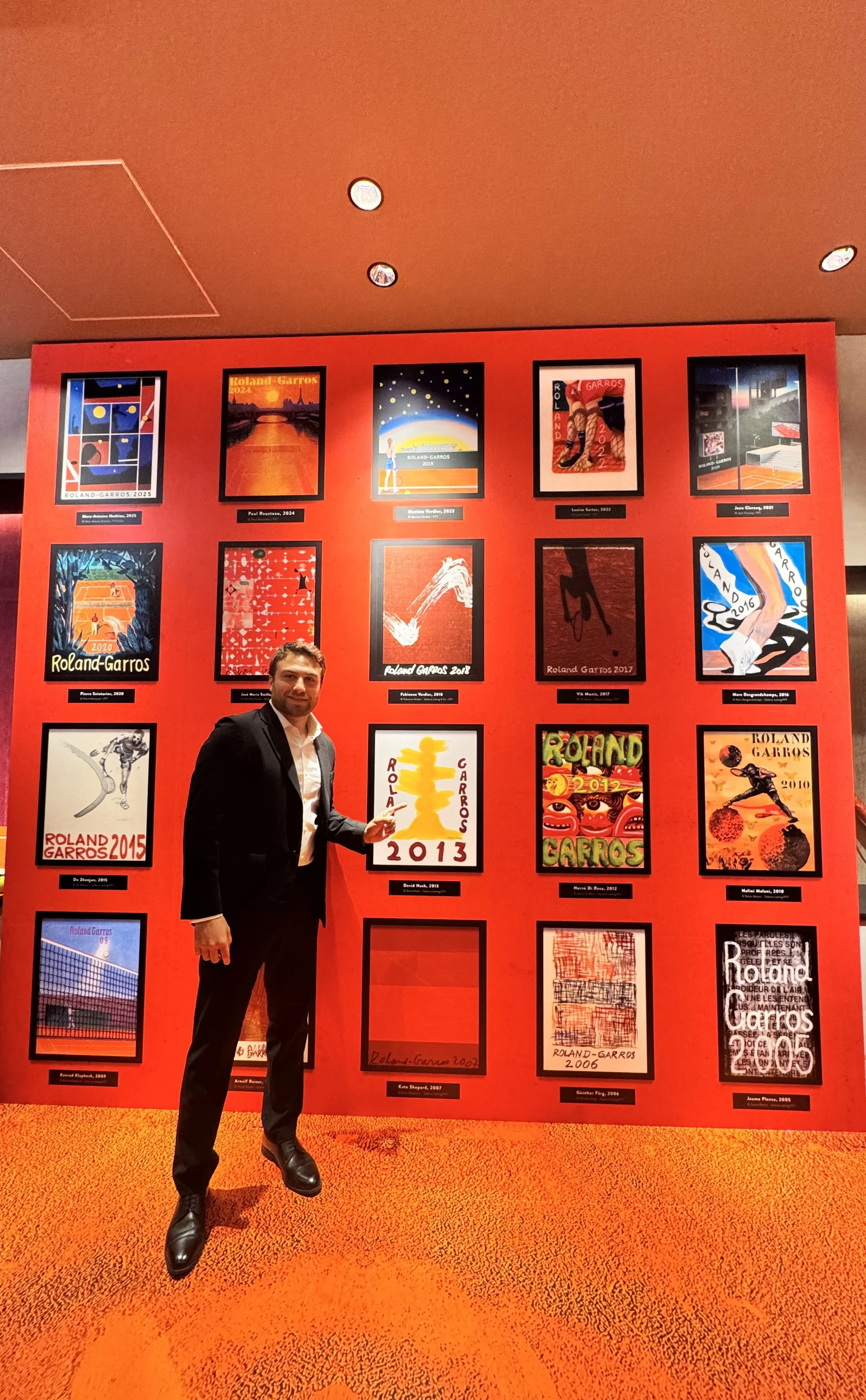 A man in a black suit poses next to a red wall displaying framed posters of the Roland-Garros tennis tournament from various years at an indoor venue.
