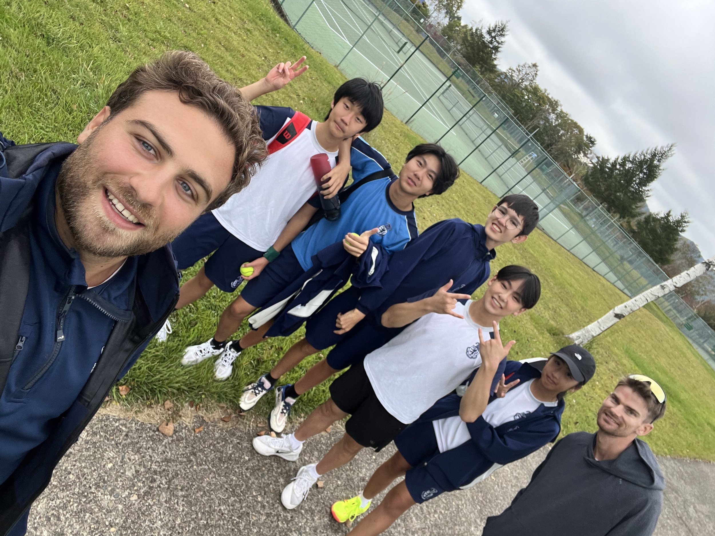 A group of six tennis players and two coaches posing for a selfie on a tennis court outdoors, with trees and a fence in the background, overcast weather.