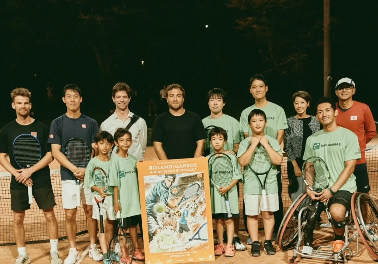 Group of young tennis players, coaches, and officials on a tennis court at night, holding tennis rackets with a poster for the Roland-Garros Junior Series event in the foreground.