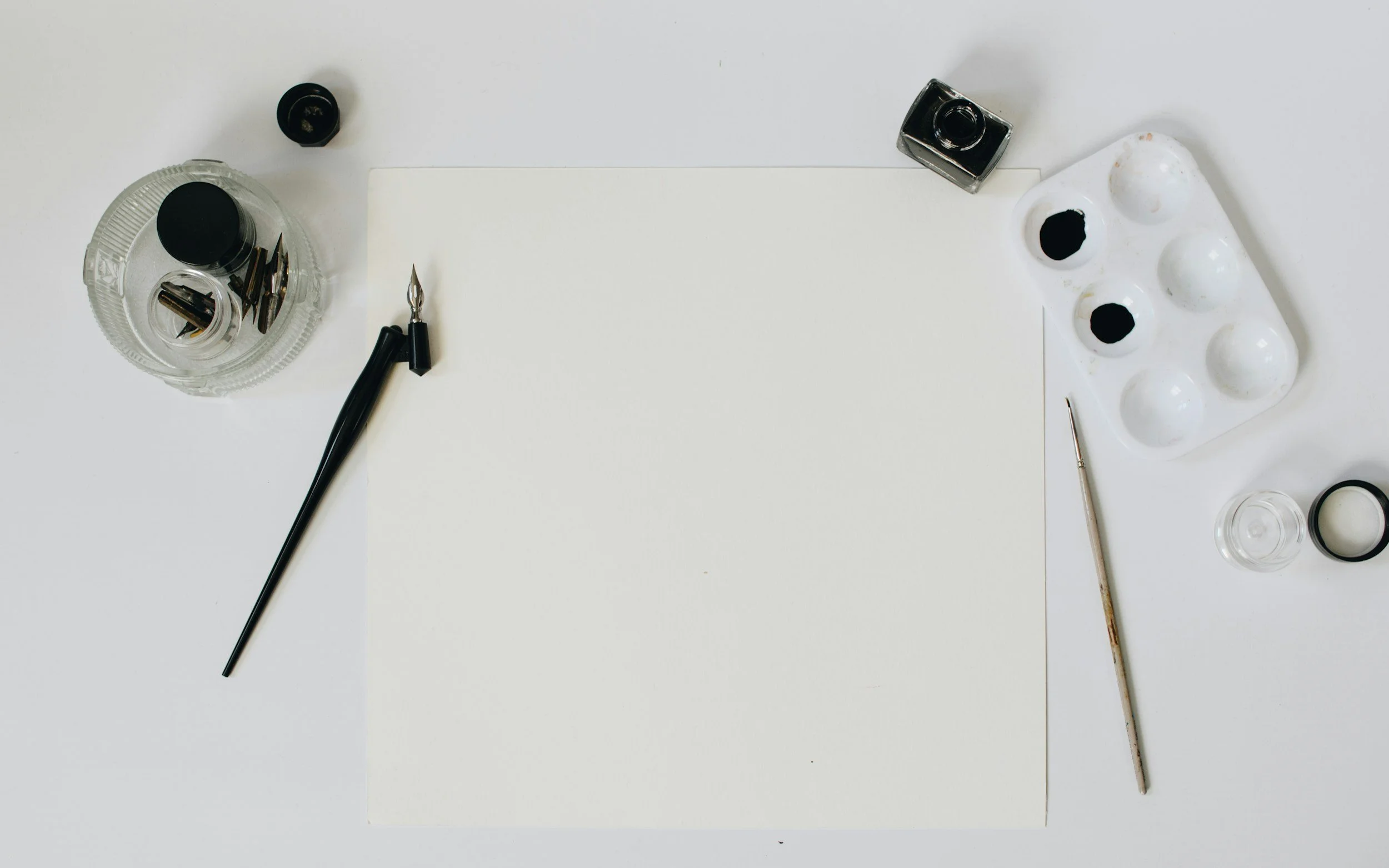 Art supplies including black ink bottles, a palette with black paint, a sheet of white paper, a calligraphy pen with nibs, an ink bottle, a thin paintbrush, and a small glass container are arranged on a white surface.