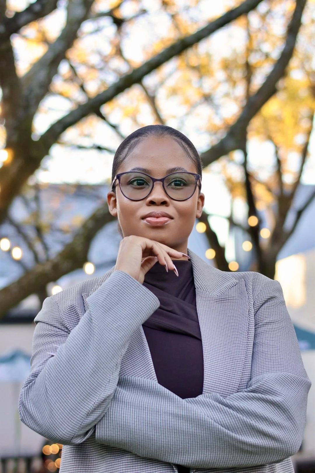 A confident woman with glasses and neatly styled hair standing outdoors in front of a tree with yellow leaves and string lights, wearing a gray blazer and dark top.
