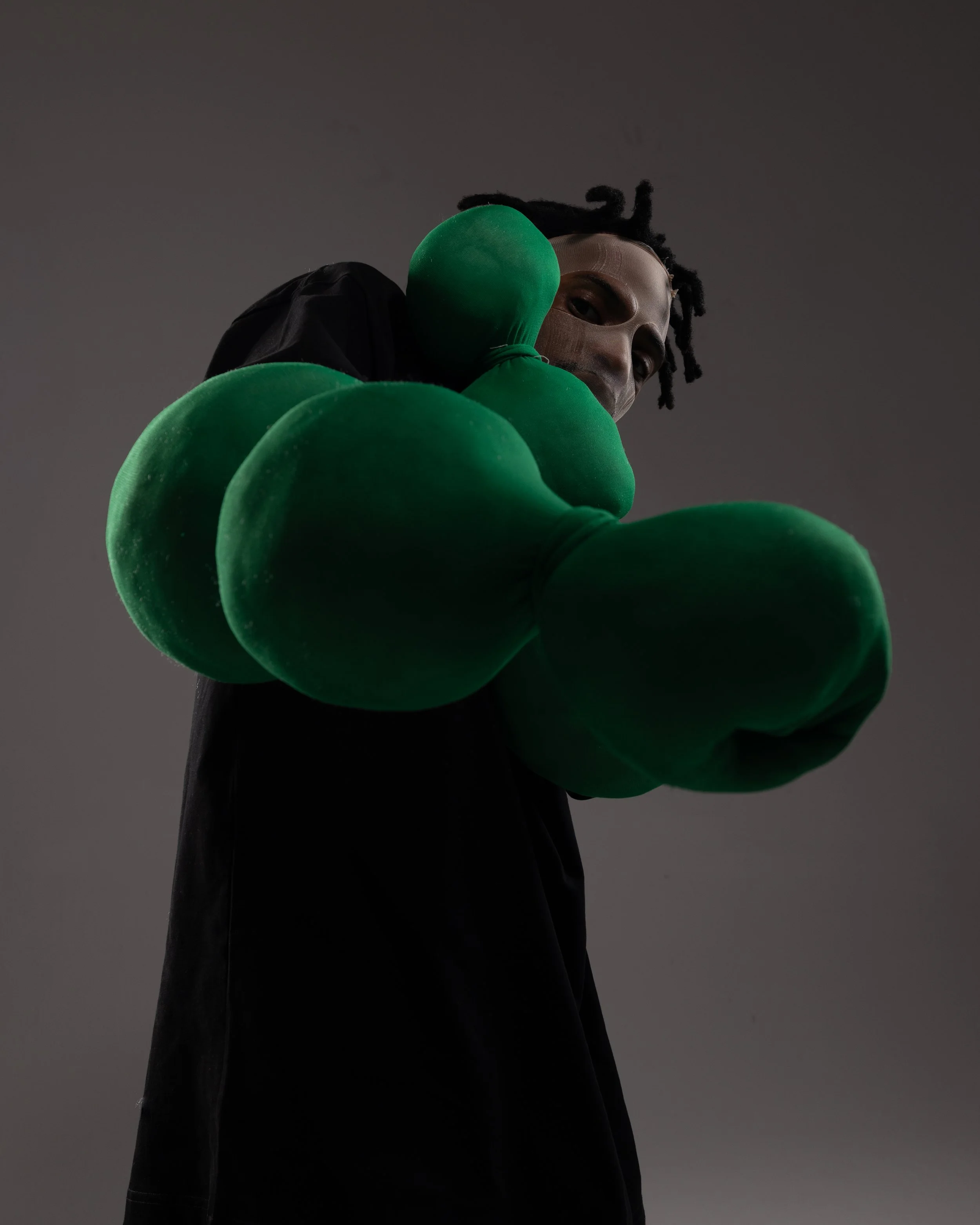 A person wearing a metallic mask and black clothing, holding or wearing large green gloves, against a dark gray background.