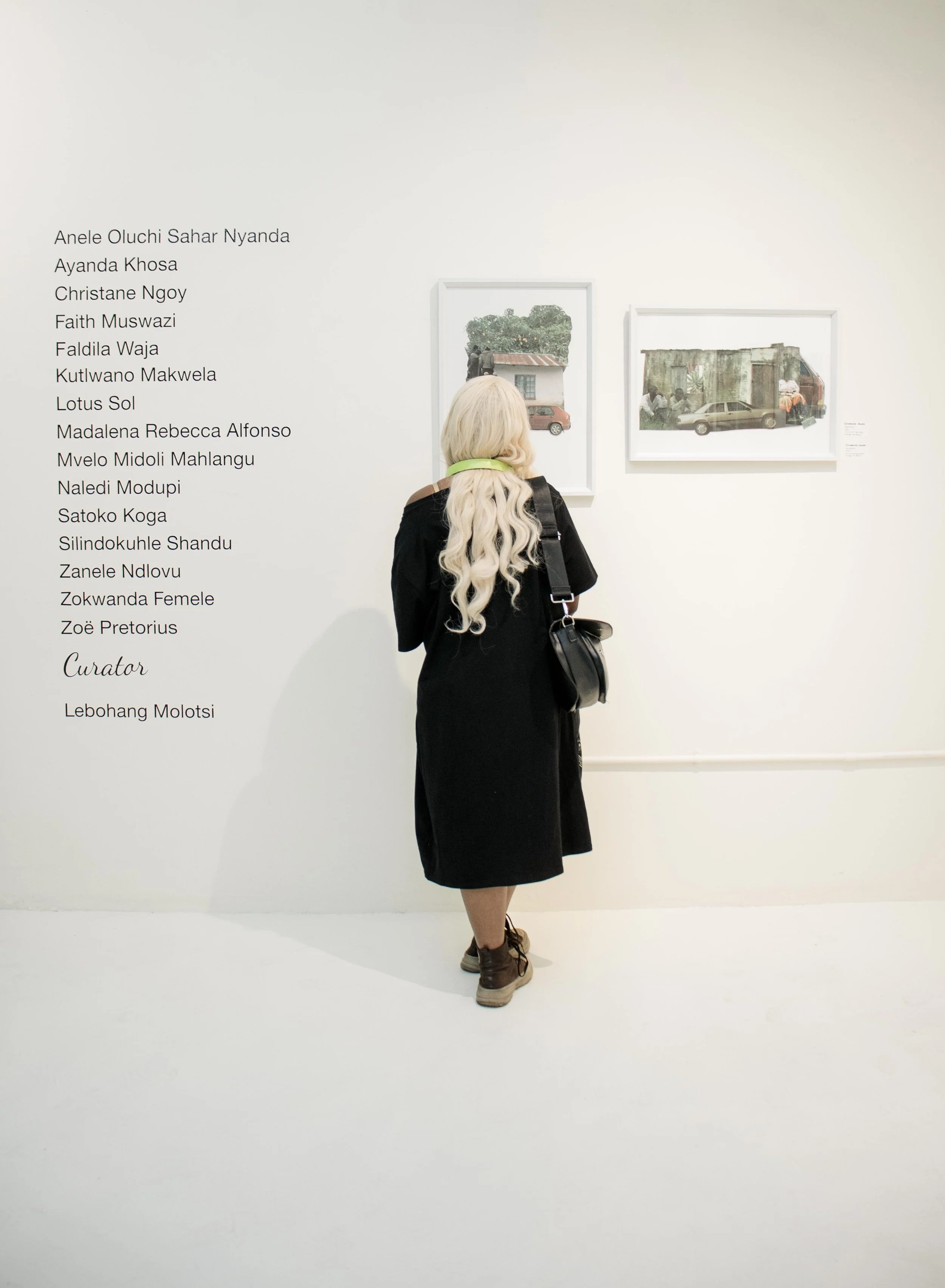A woman with long blonde hair, wearing a black coat, brown shoes, and a green headband, stands with her back to the camera, looking at framed photographs on a gallery wall.