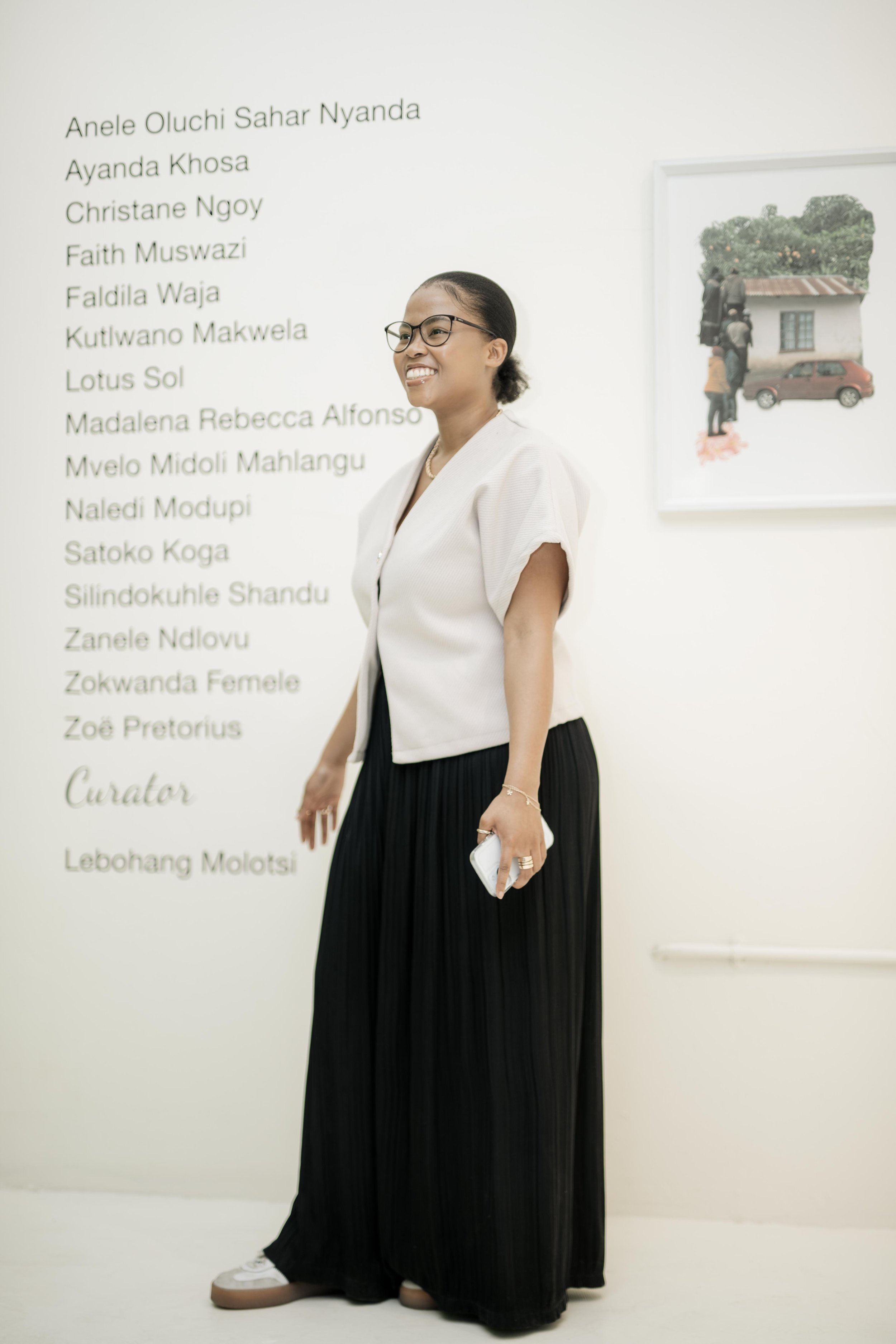 A woman wearing glasses, a white blazer, and a long black skirt, standing inside an art gallery or museum with a white wall, looking to the side and smiling, holding a phone in one hand.