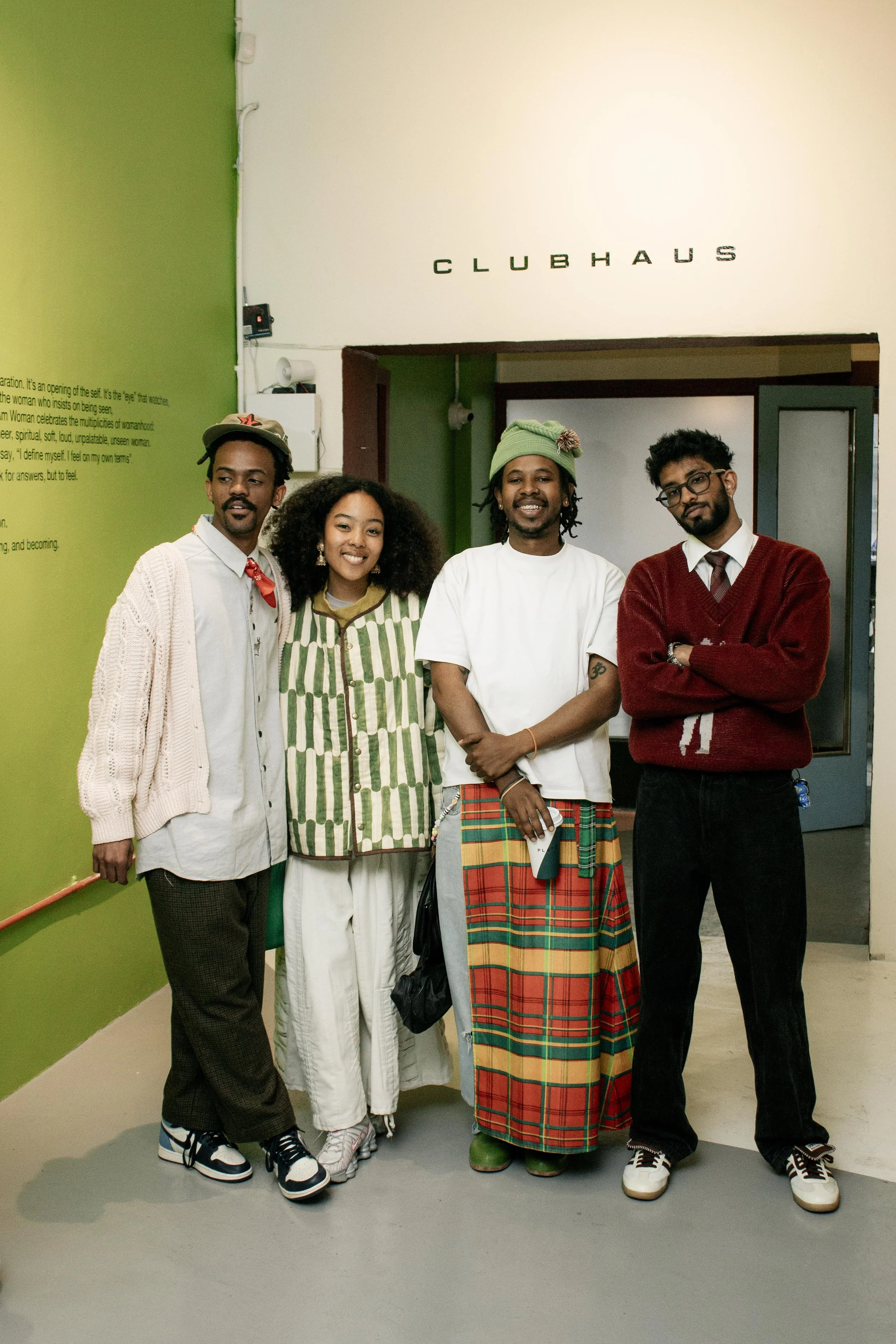 A group of five diverse people, consisting of three men and two women, standing together indoors in front of a wall with the word 'CLUBHAUS' on it. They are smiling and dressed in a variety of casual and colorful clothing, with one person wearing a g