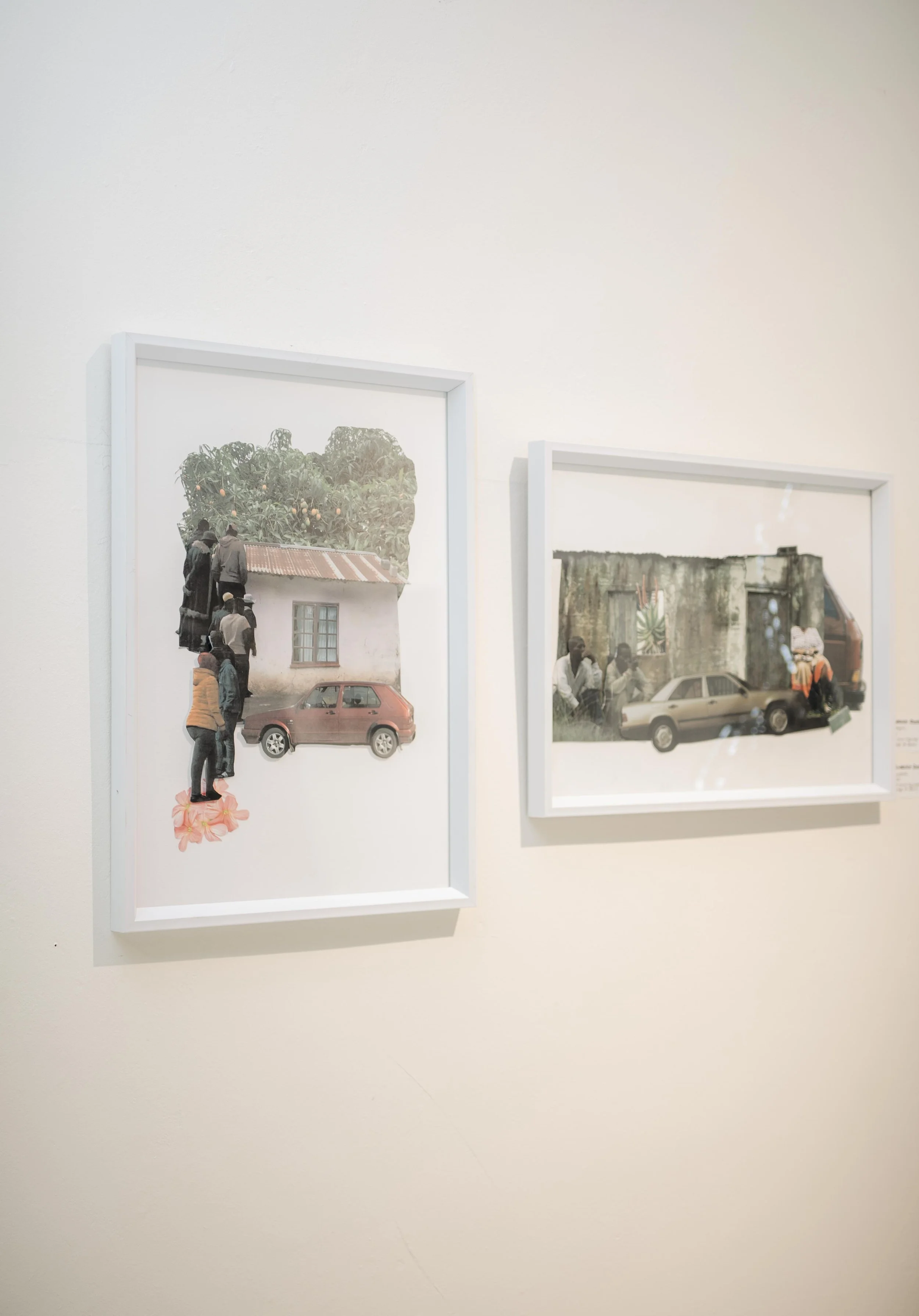 Two framed photographs hanging on a white gallery wall. The left photo depicts a house, a vintage car, and people with a large tree in the background. The right photo shows people standing outside in front of a rustic building with a vintage car.