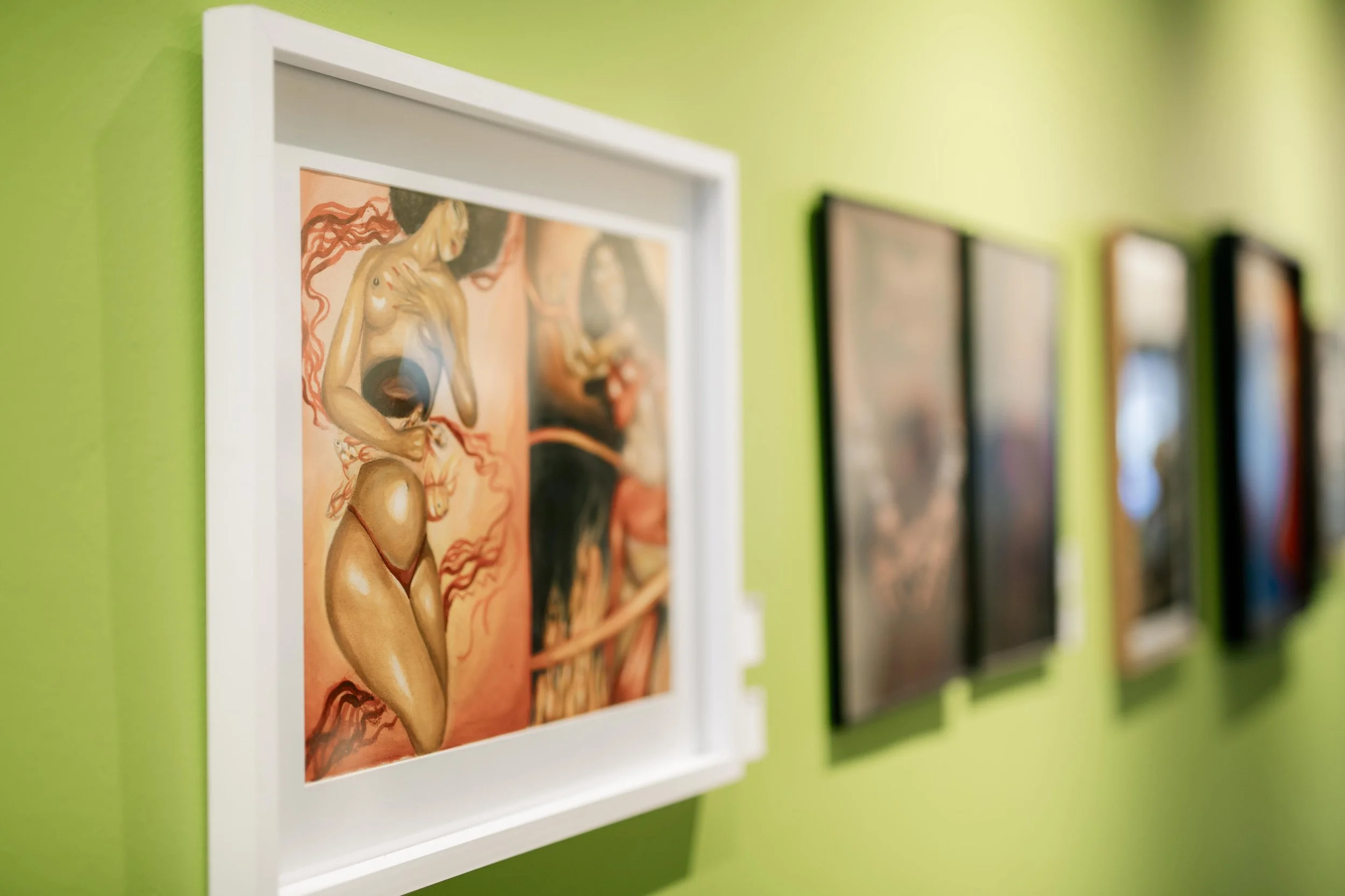 Close-up of framed artwork depicting a topless woman with long flowing hair, set against a green wall, with other artworks partially visible in the background.