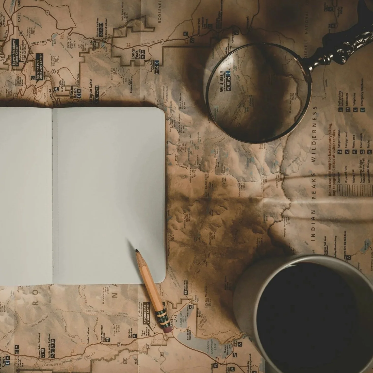 A top-down view of a desk with a map of Wilderness, a pencil, a gray notebook, and a mug filled with a dark beverage. The map shows various trails and points of interest in a wilderness area.