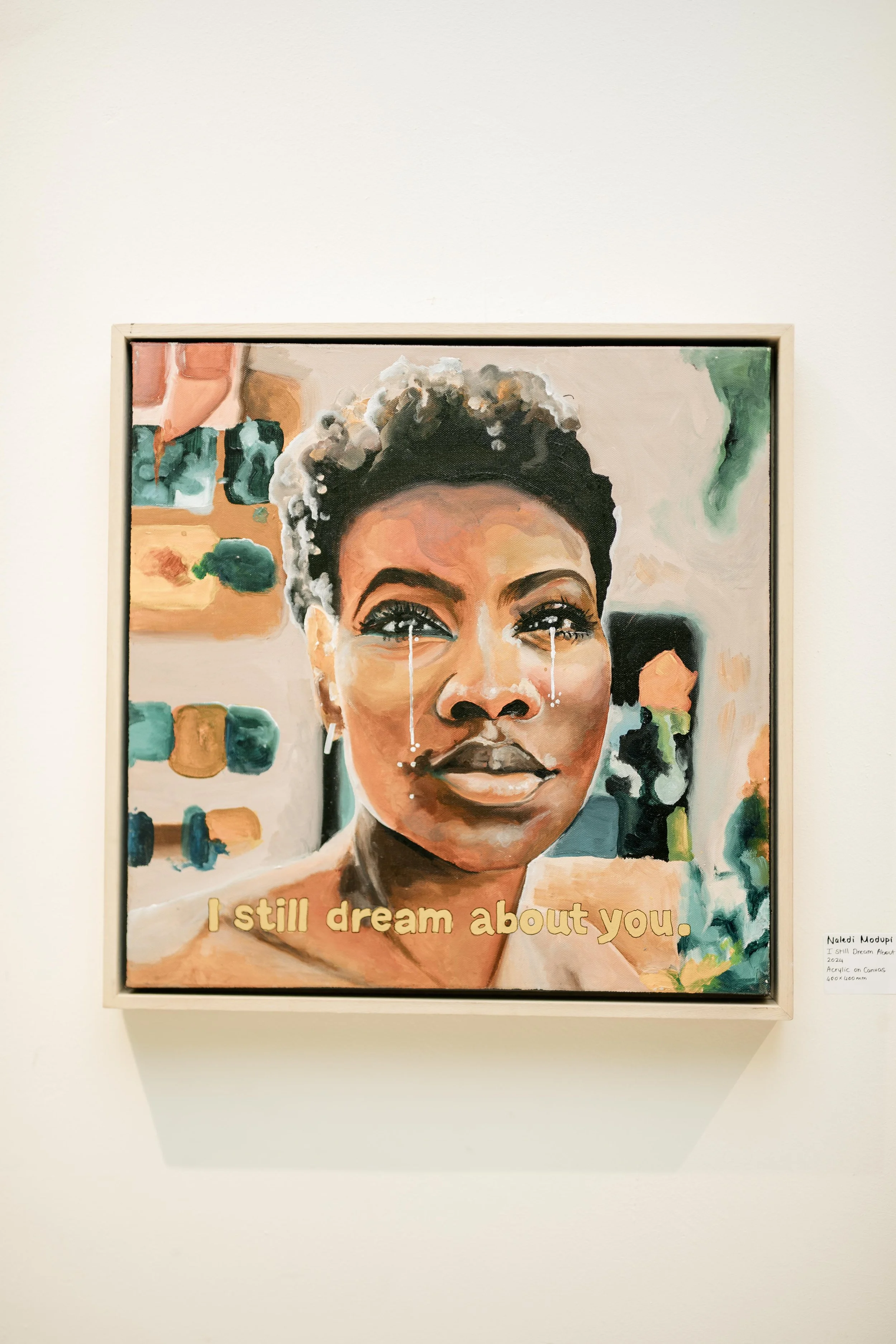 A colorful painting of a woman with tears streaming down her face, with a quote at the bottom that reads 'I still dream about you.' The artist's name 'Naled Molupi' and details about the artwork are written on a small label at the bottom right corner