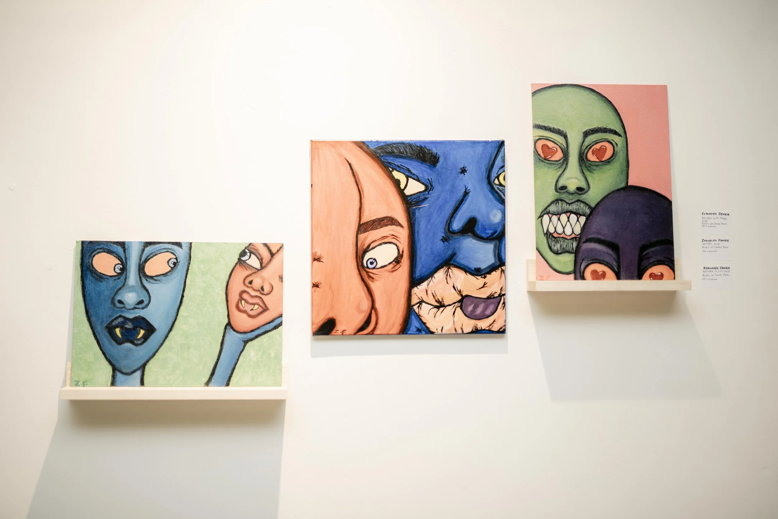 Colorful paintings of expressive faces with exaggerated features and bold outlines, displayed on a white gallery wall.