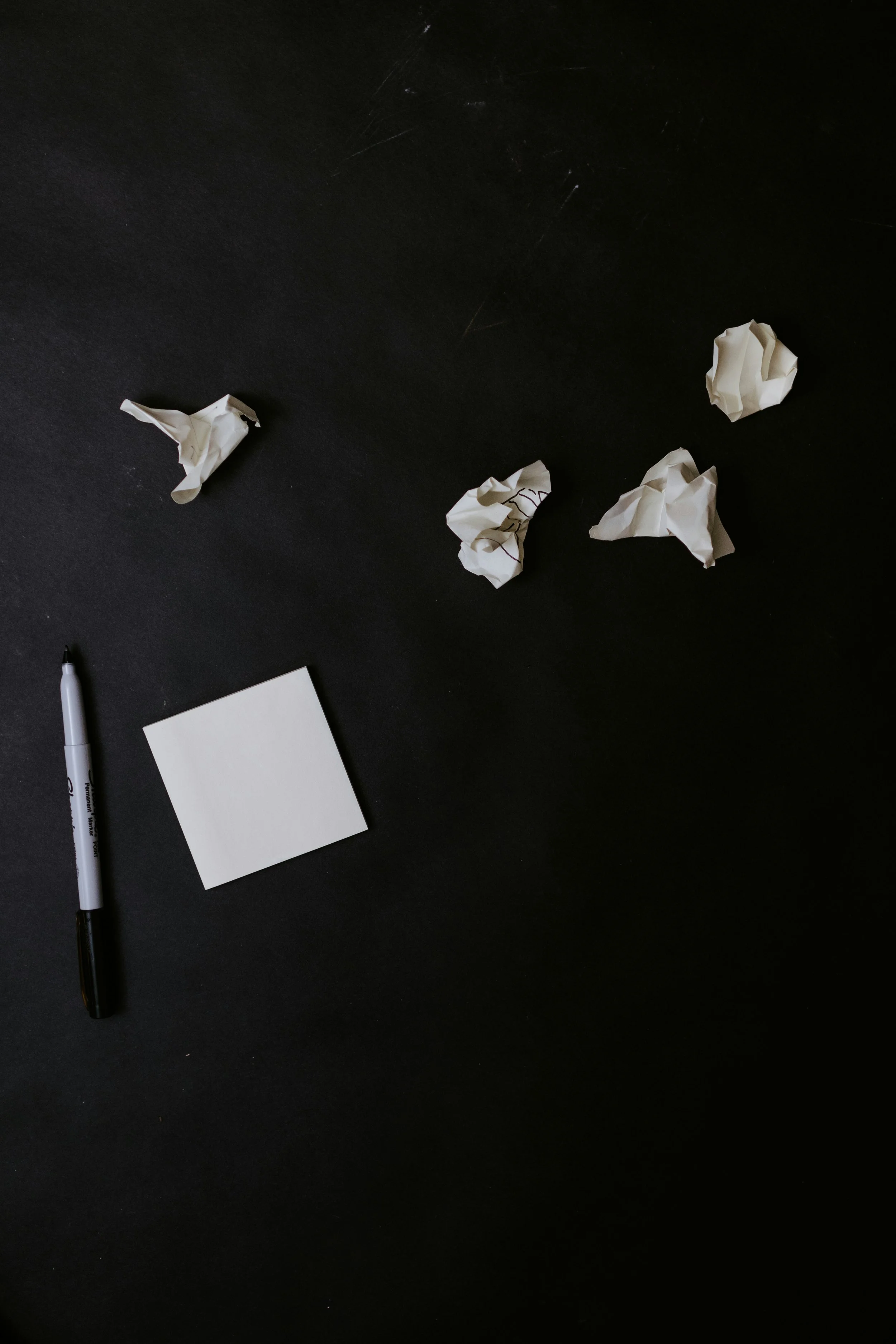 A black surface with crumpled white paper balls scattered across it, a black and white marker pen, and a small blank white square paper.